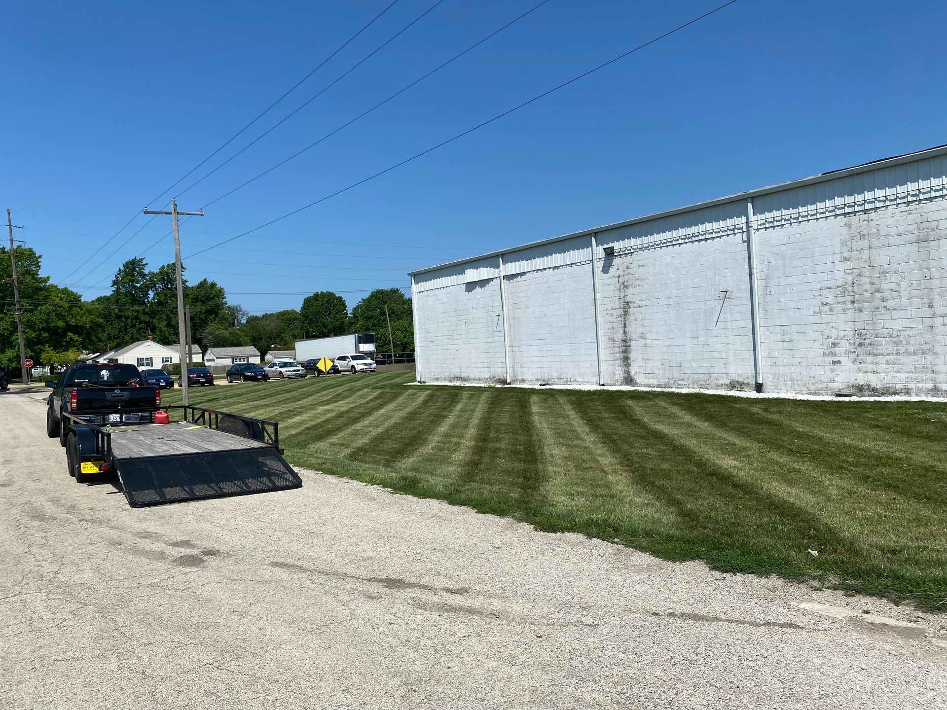 Black pickup towing a flatbed trailer on a gravel road beside a white industrial building.