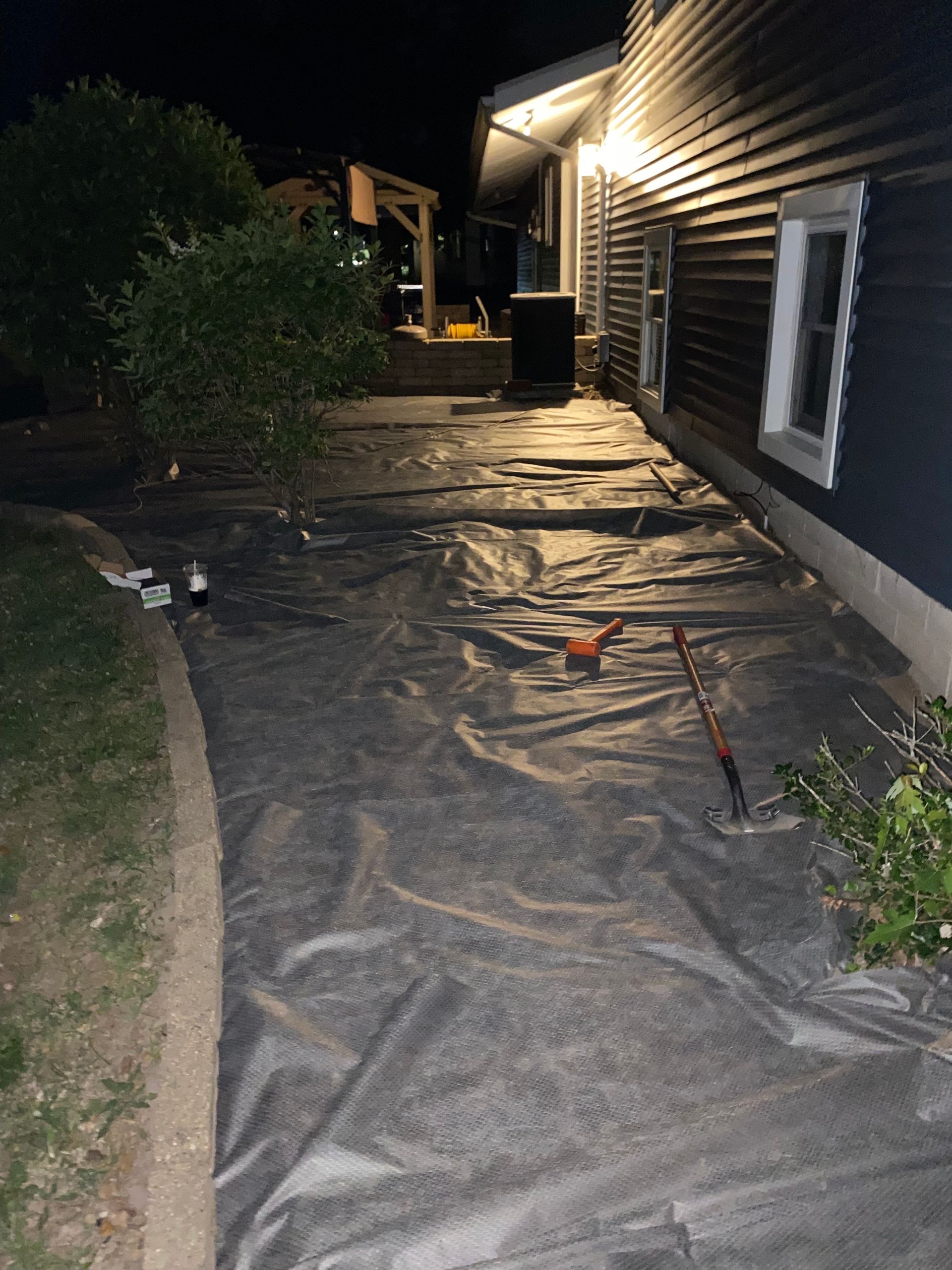 Nighttime backyard pathway covered with black landscape fabric beside a house and bushes
