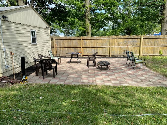 Backyard patio with brick pavers, outdoor chairs, fire pit, and wooden fence beside a house.