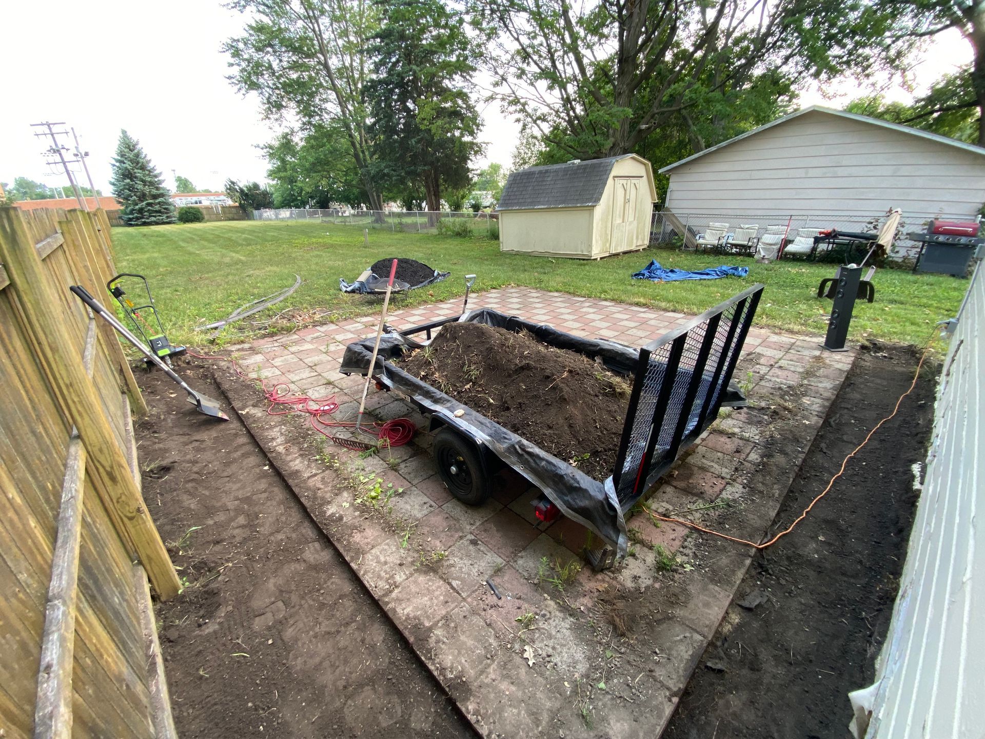 Soil-filled trailer parked in a backyard next to a fenced garden area and shed