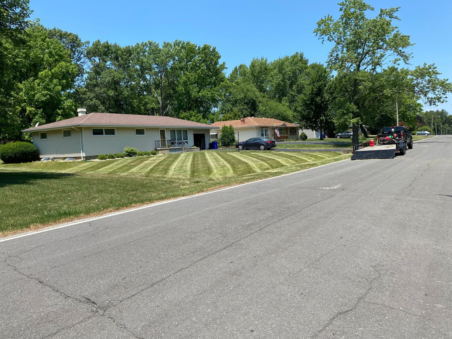 Quiet suburban street with single-story houses, green lawns, trees, and parked cars on a sunny day