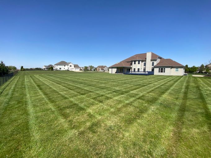 Wide mowed grassy field under blue sky, with large house and distant homes in the background.