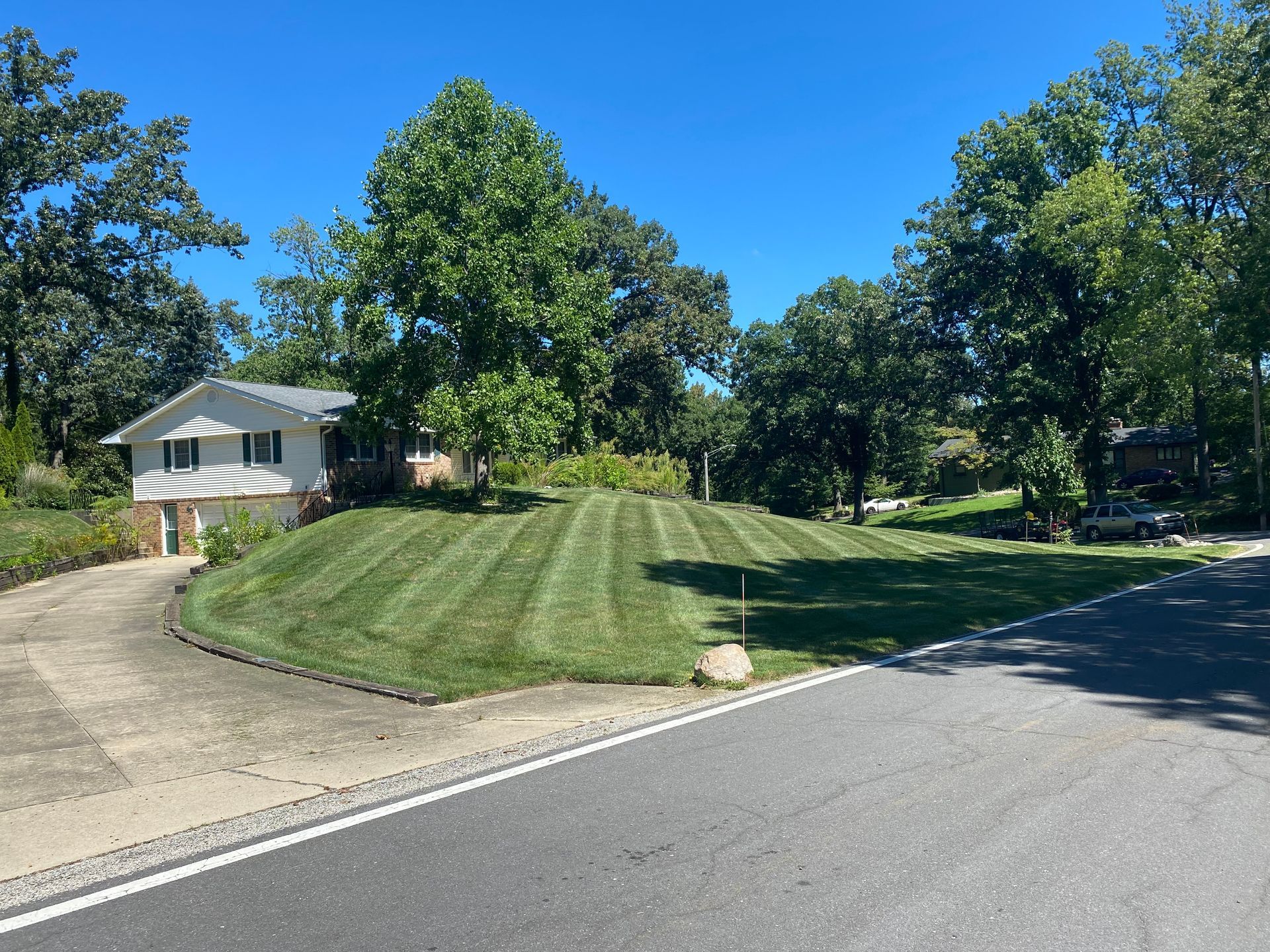 Suburban corner with a mowed grassy lawn, trees, a white house, and a paved street under a clear blue sky