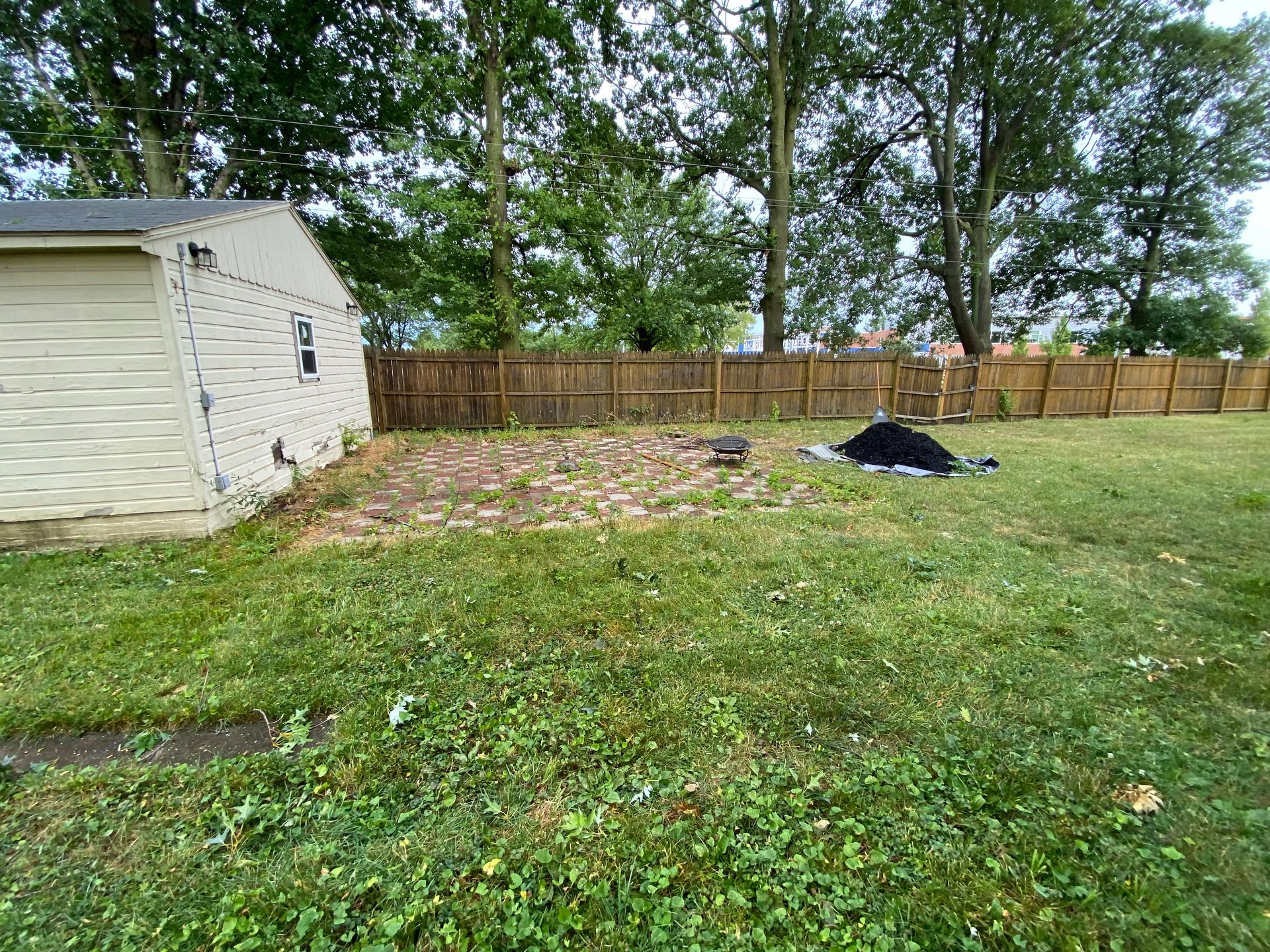 Backyard with a small shed, patchy grass, wooden fence, and a pile of debris near the center.