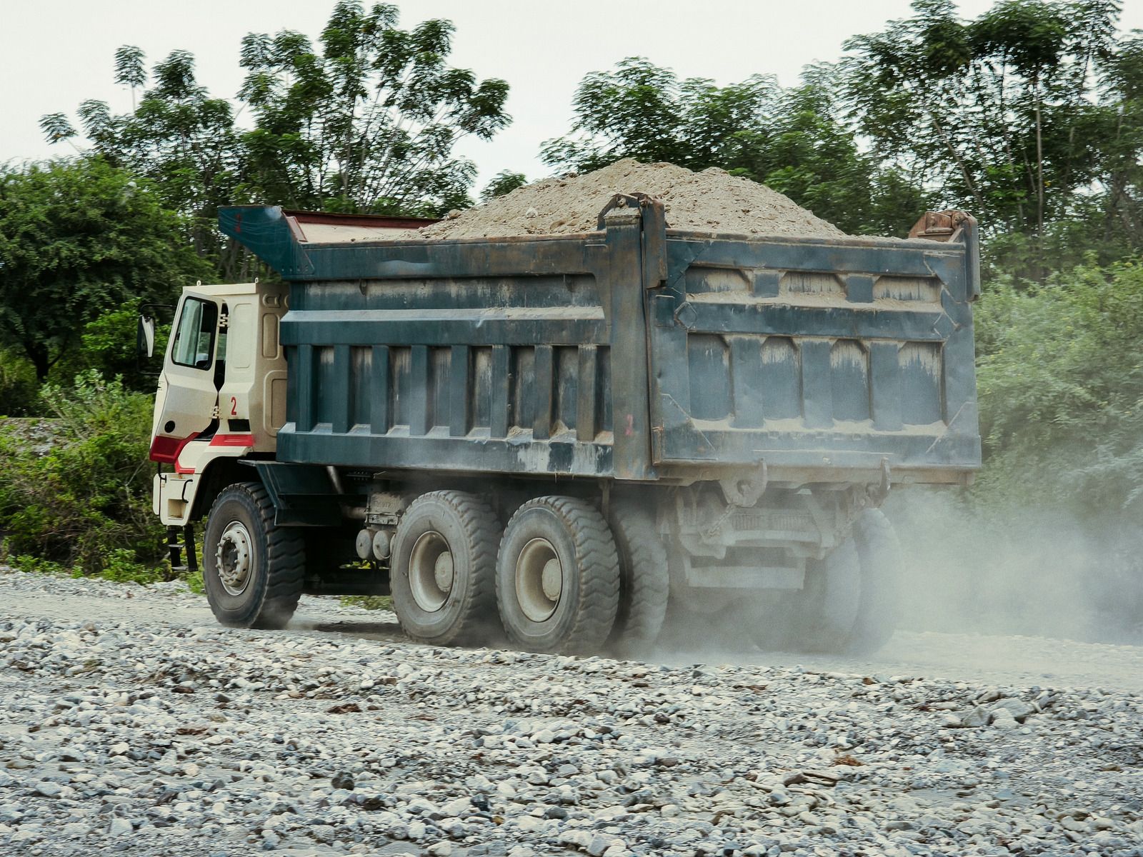 Dump truck loaded with gravel driving on a dusty gravel road near trees