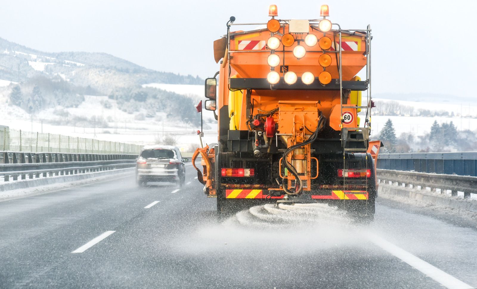 Orange road maintenance truck spraying water on a snowy highway, with a car behind it.