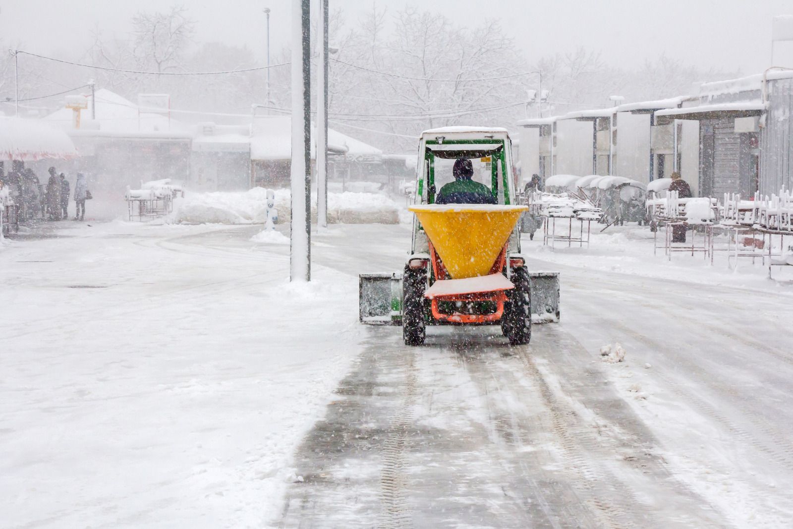 Snowplow clearing a snowy street, with a driver in a green cab and yellow front blade.