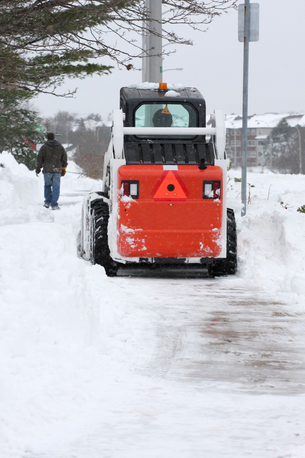 Orange snow-clearing vehicle on a snowy sidewalk with a person walking nearby