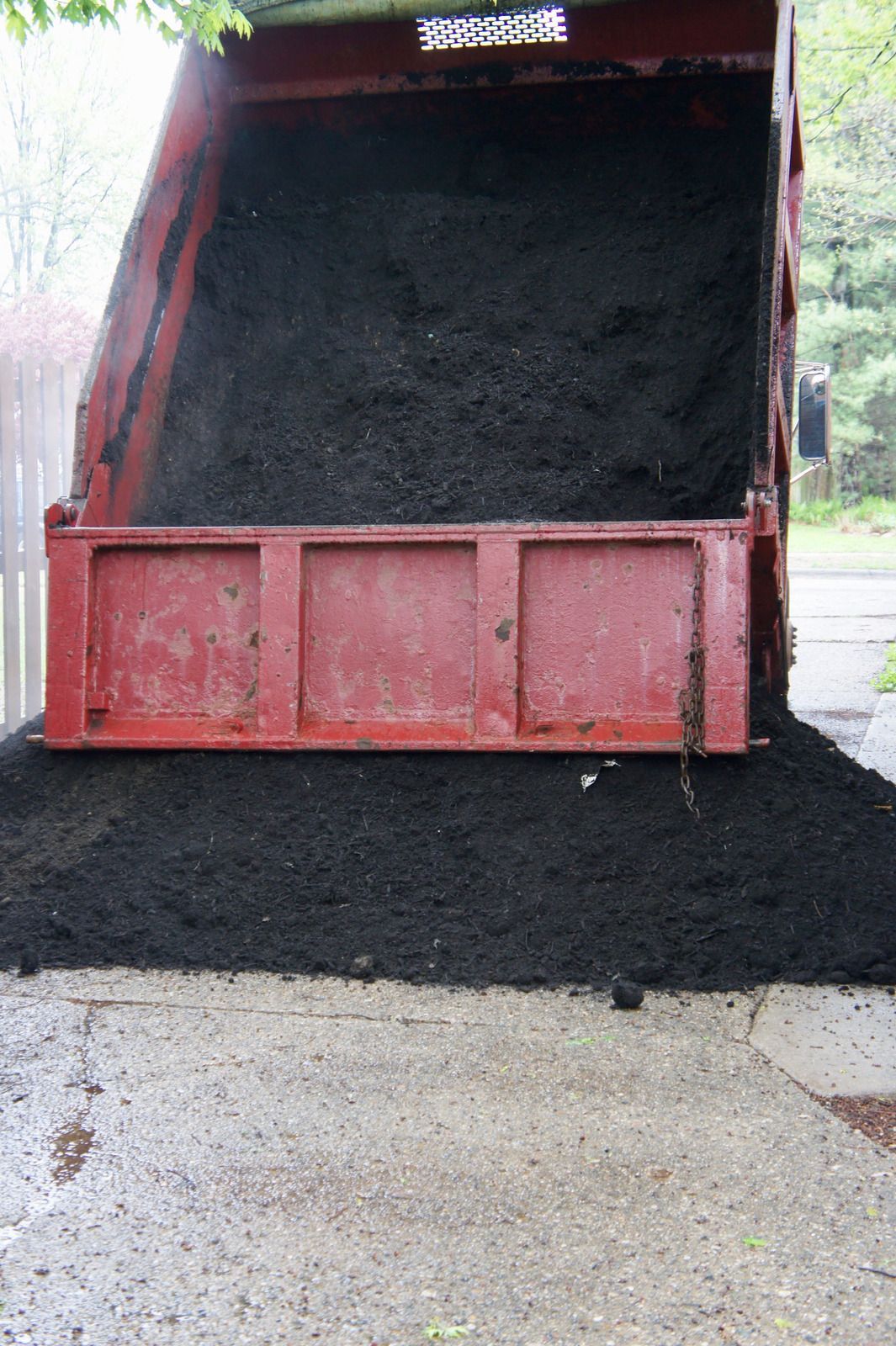 Open red dump truck bed spilling black soil onto a concrete driveway