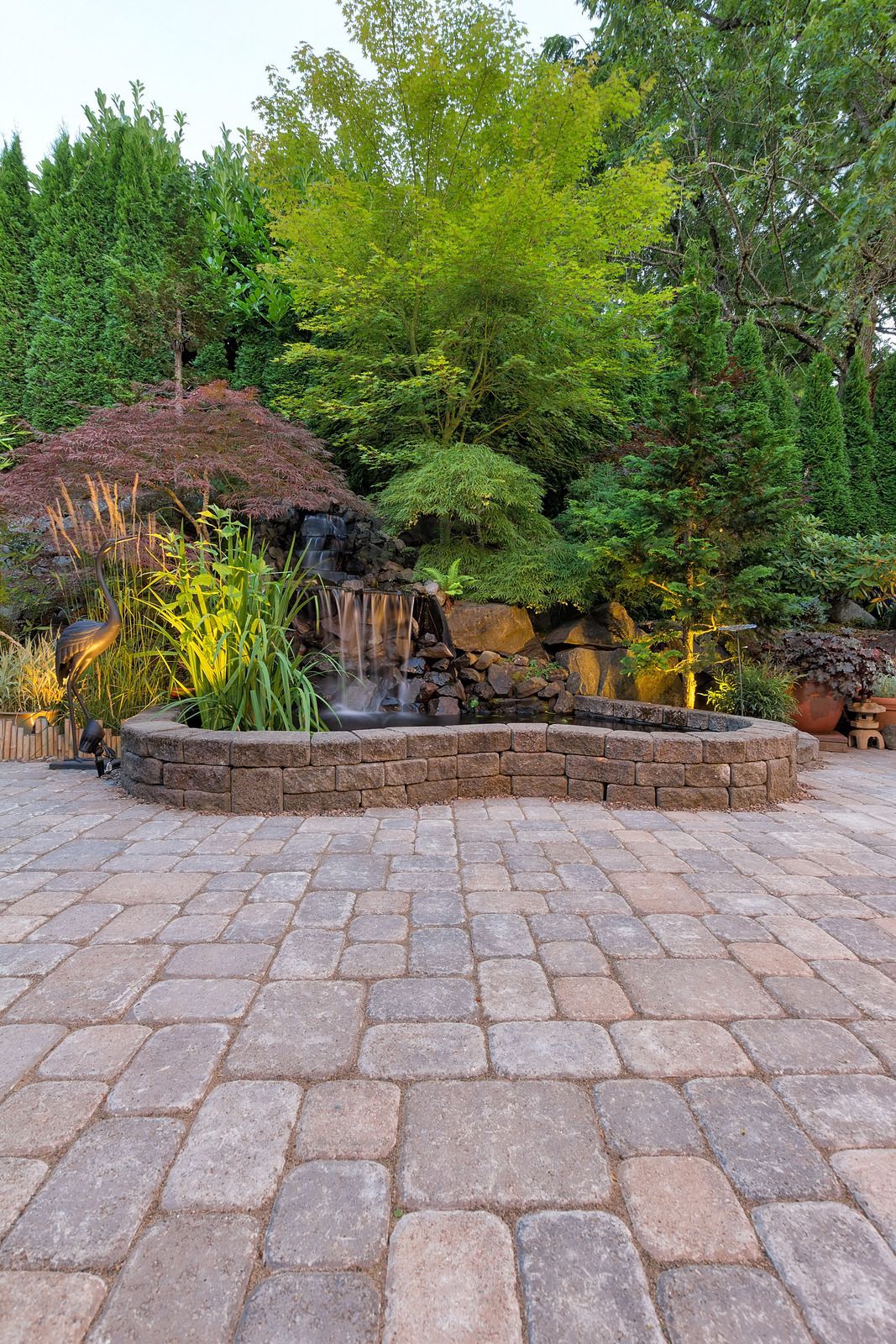 Stone patio with a small cascading waterfall, lush greenery, and warm garden lights at dusk