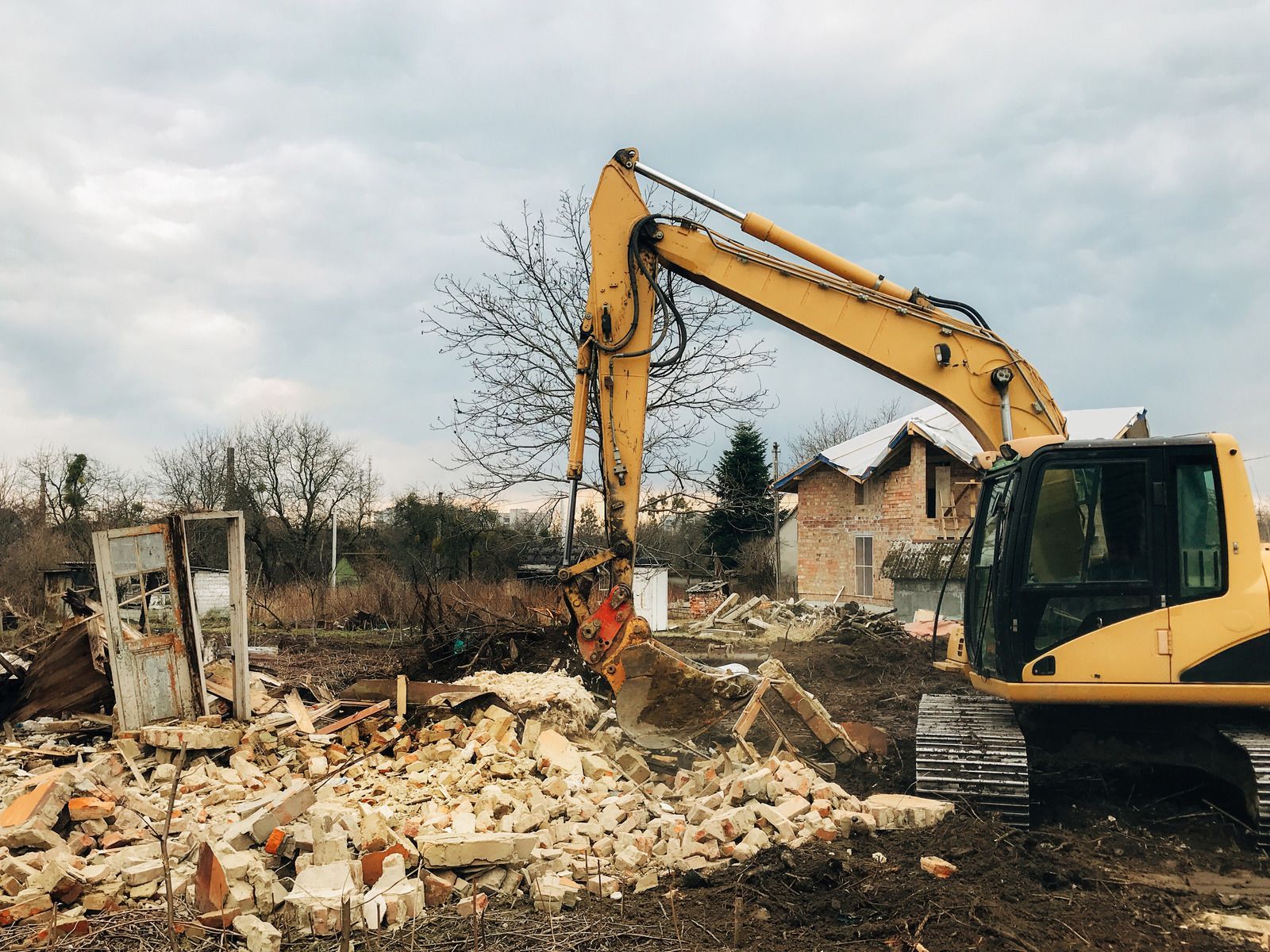 Yellow excavator demolishing a rubble-strewn building site under a cloudy sky