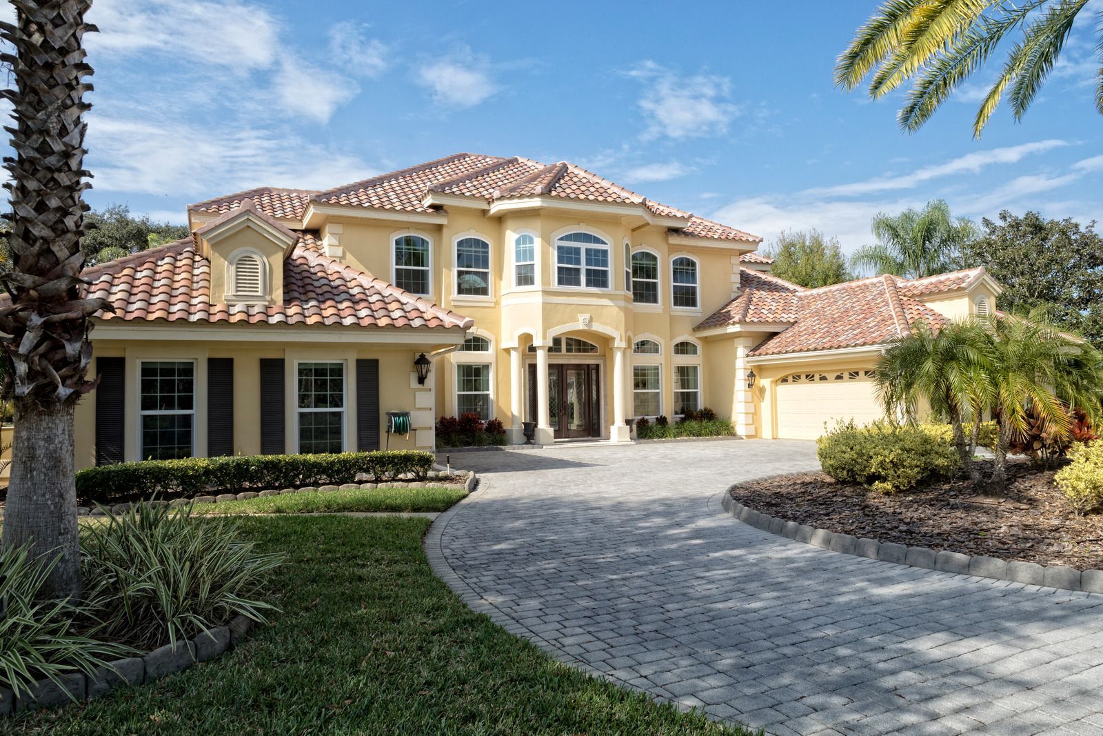 Luxury beige stucco house with red tile roof and curved driveway under palm trees