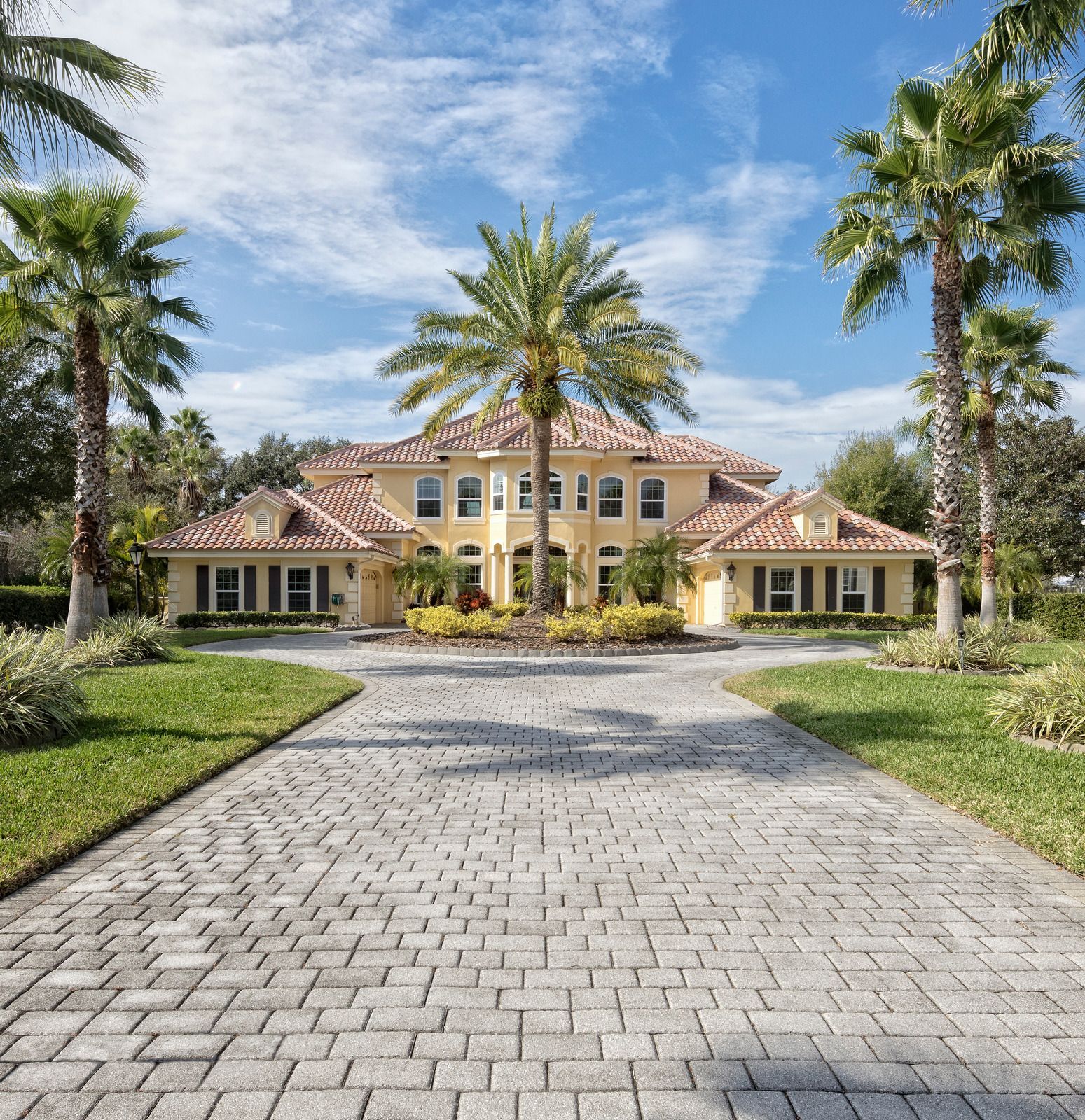 Large Mediterranean-style villa with a brick driveway, palm trees, and manicured landscaping under a blue sky