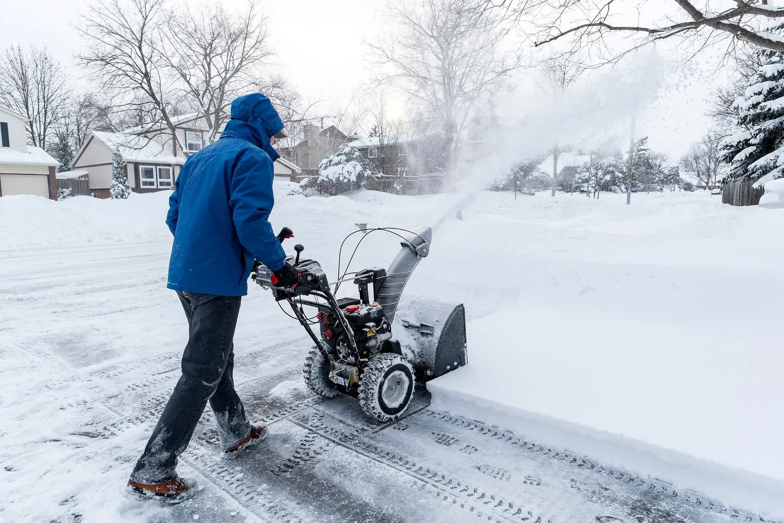 Person in a blue jacket clearing snow with a snowblower on a snowy residential street