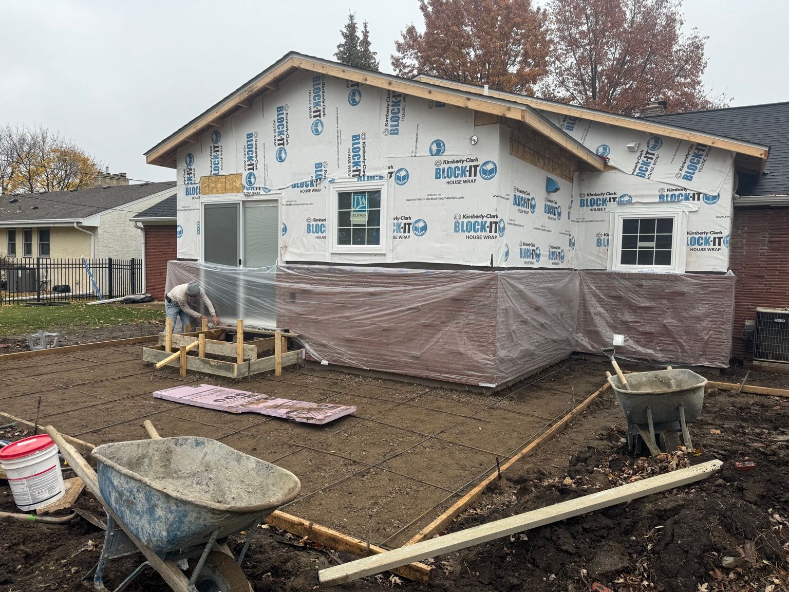 House under construction wrapped in housewrap, with mud, wheelbarrow, and building materials in the yard
