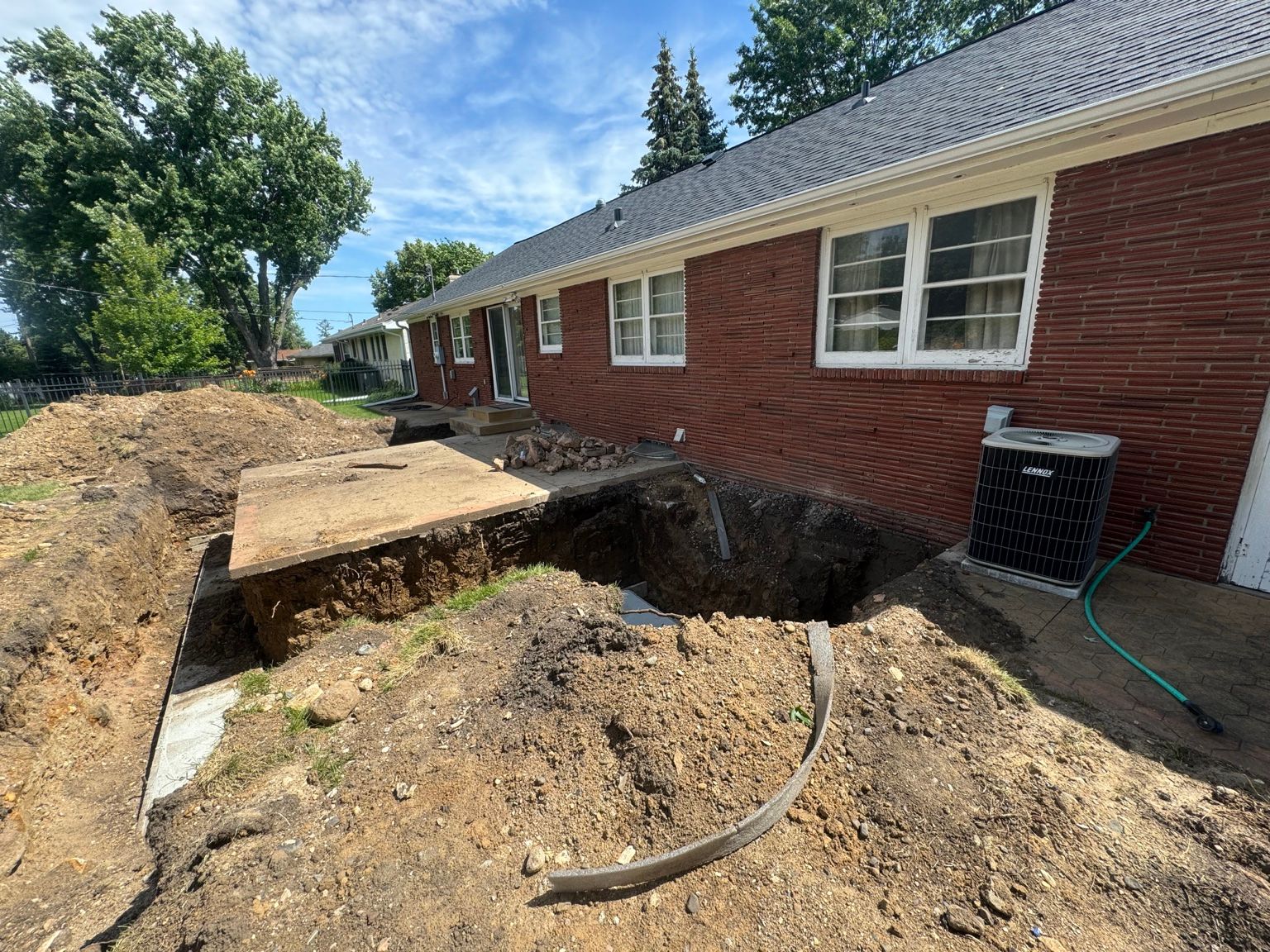Backyard excavation beside a red brick house, with a collapsed dirt trench and wooden planks over the hole