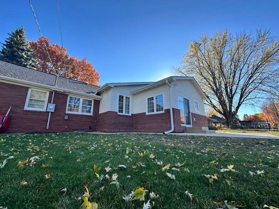 Single-story red-brick house with white siding and a large leafless tree in a grassy yard under a blue sky