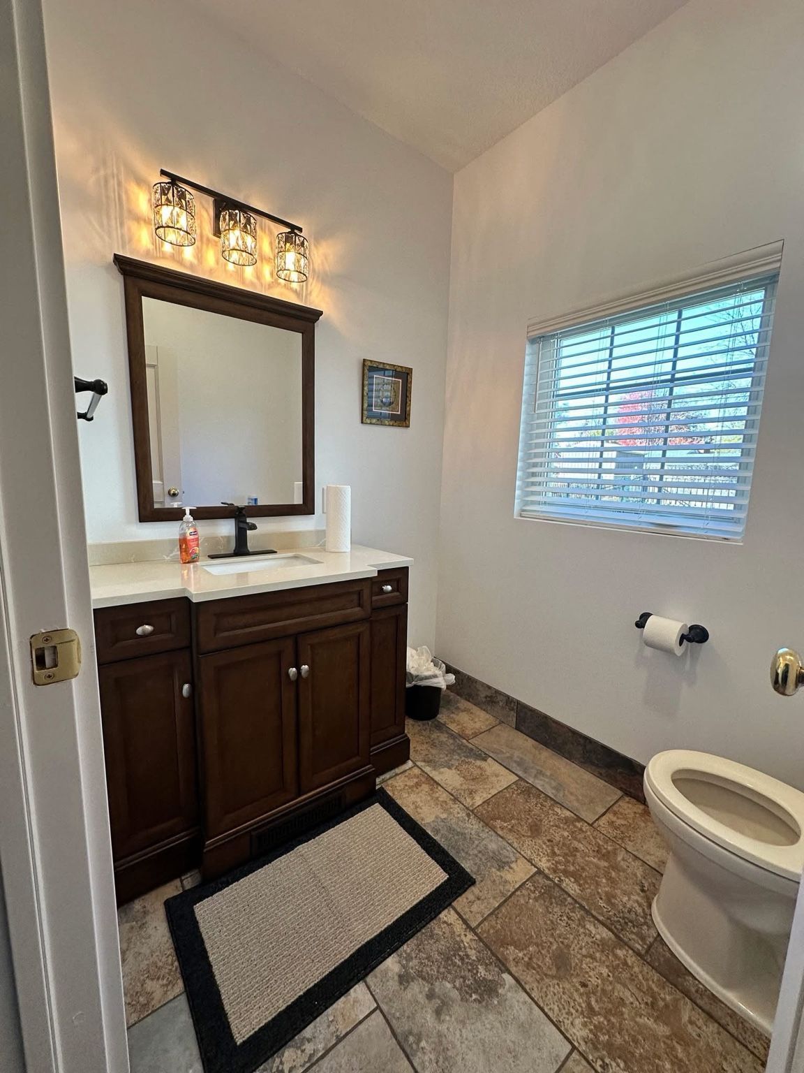 Small bathroom with dark vanity, mirror lights, window blinds, and a toilet on a tiled floor.