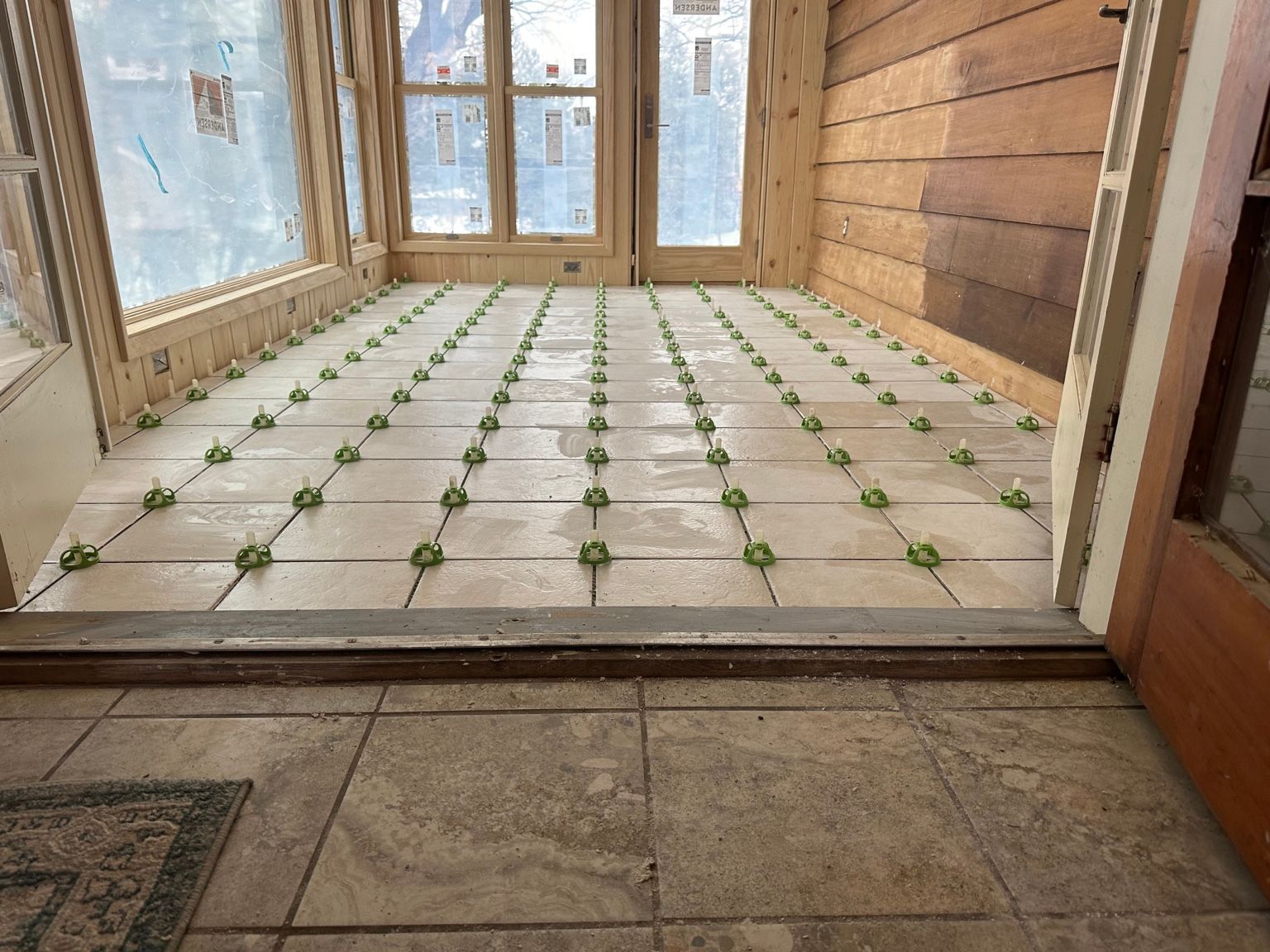 Room with tiled floor and many small green plants arranged in a grid near a windowed wall.