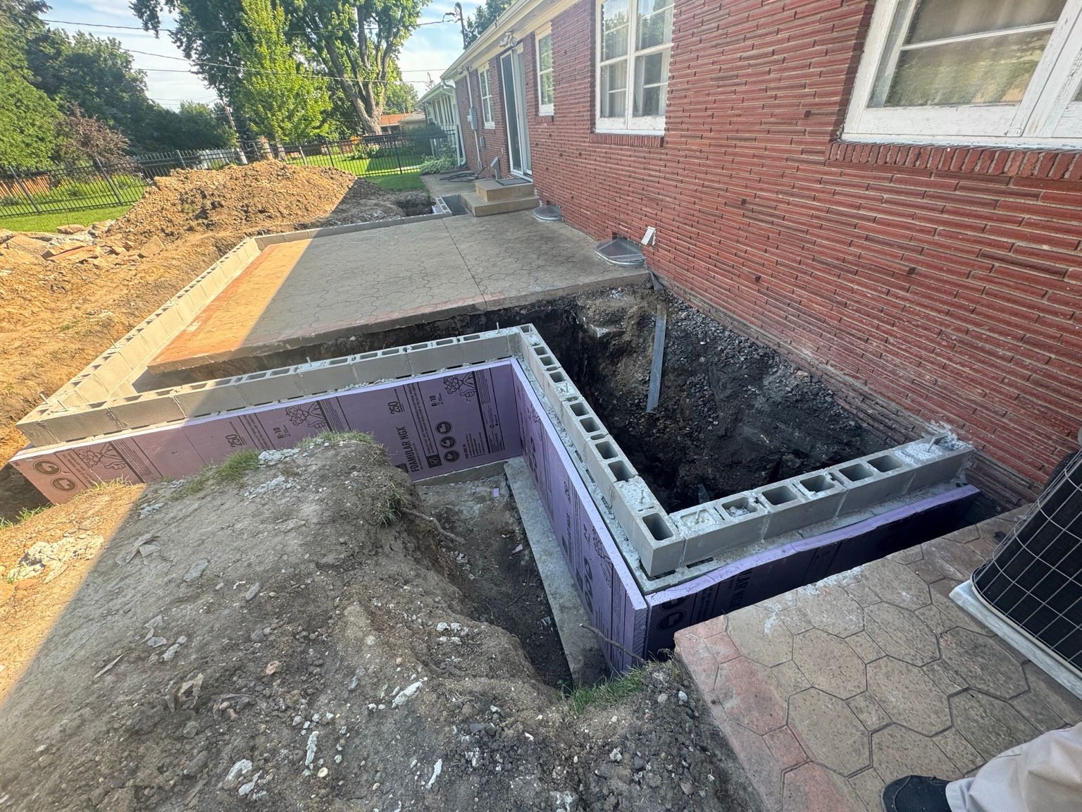 Outdoor construction trench beside a brick building, with cinder blocks and a partially dug foundation.