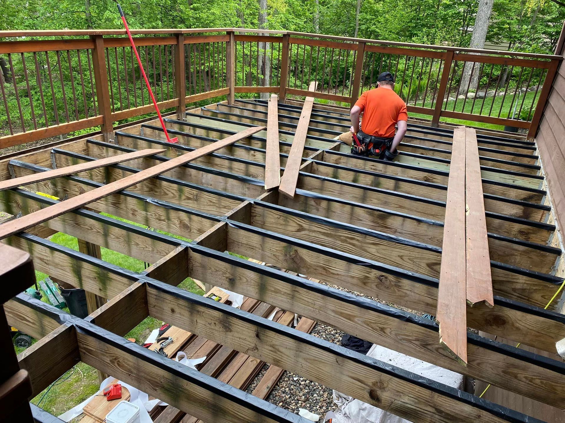 Wooden deck frame under construction with a worker installing joists on a wooded backyard deck.