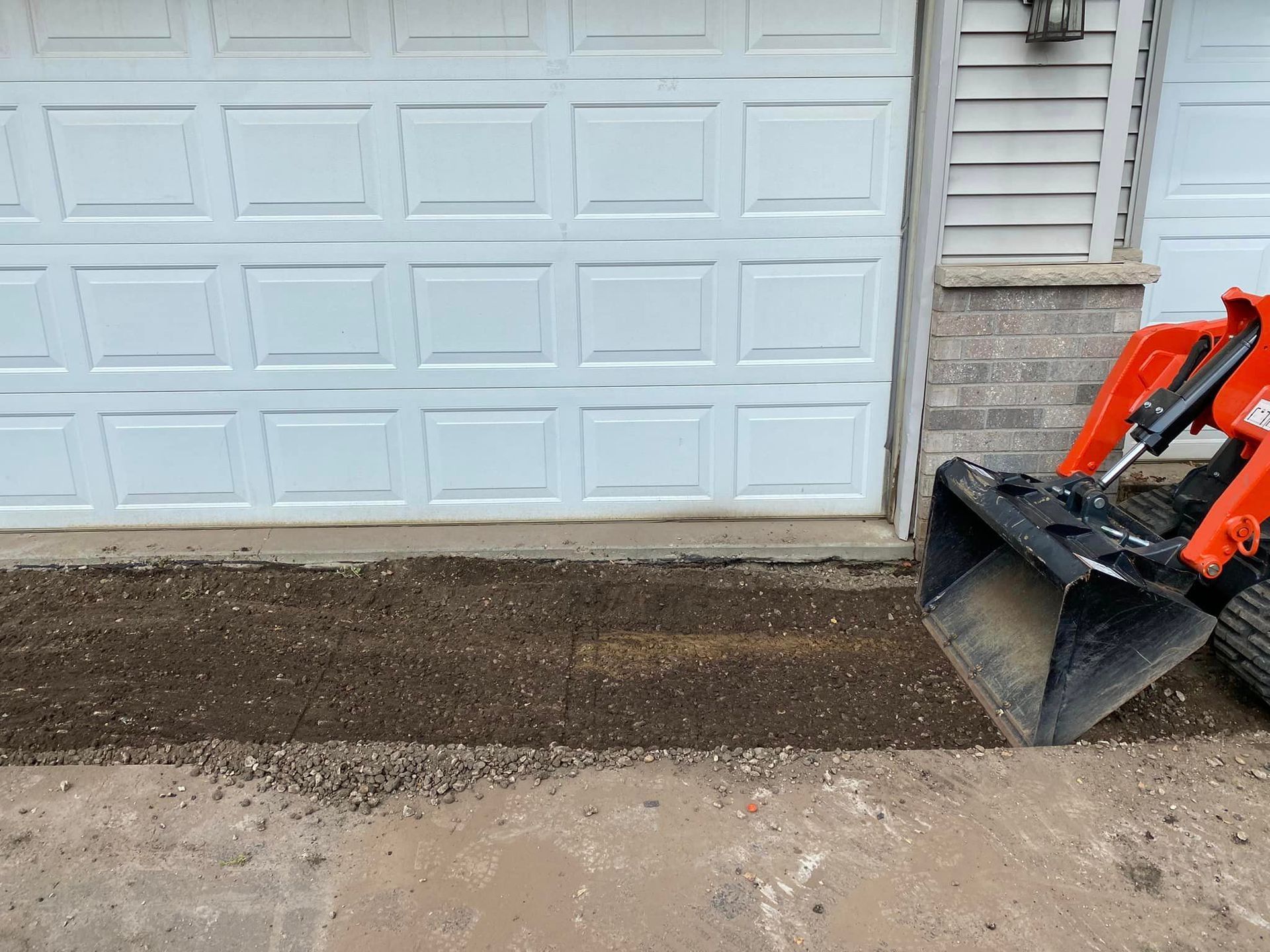 Fresh dirt laid along a garage driveway beside a white door, with an orange excavator bucket at right