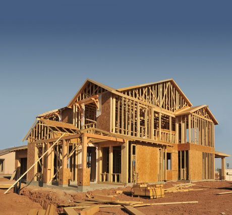 Partially built wooden house frame on a dirt lot under a clear blue sky