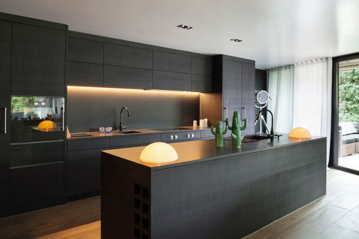 Modern black kitchen with island, built-in appliances, under-cabinet lighting, and large window to the right