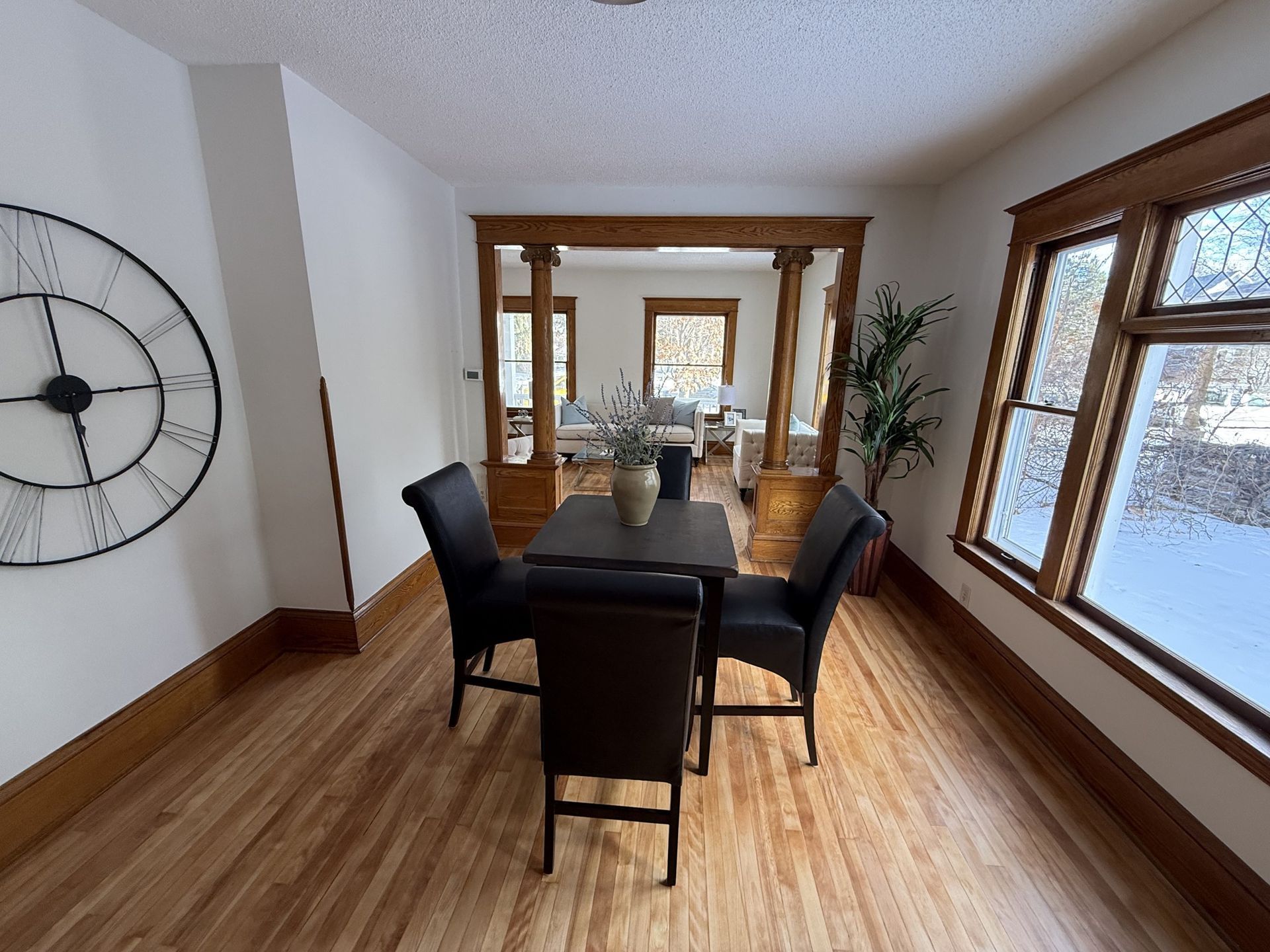 Dining room with dark table and chairs, wooden trim, wall clock, and large windows letting in snowlight
