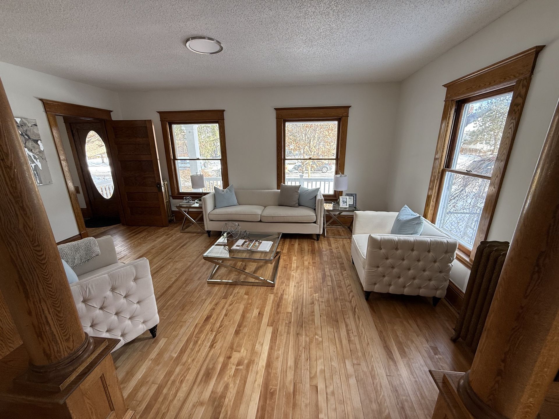 Bright living room with wood floors, beige sofa, white armchairs, and large windows.