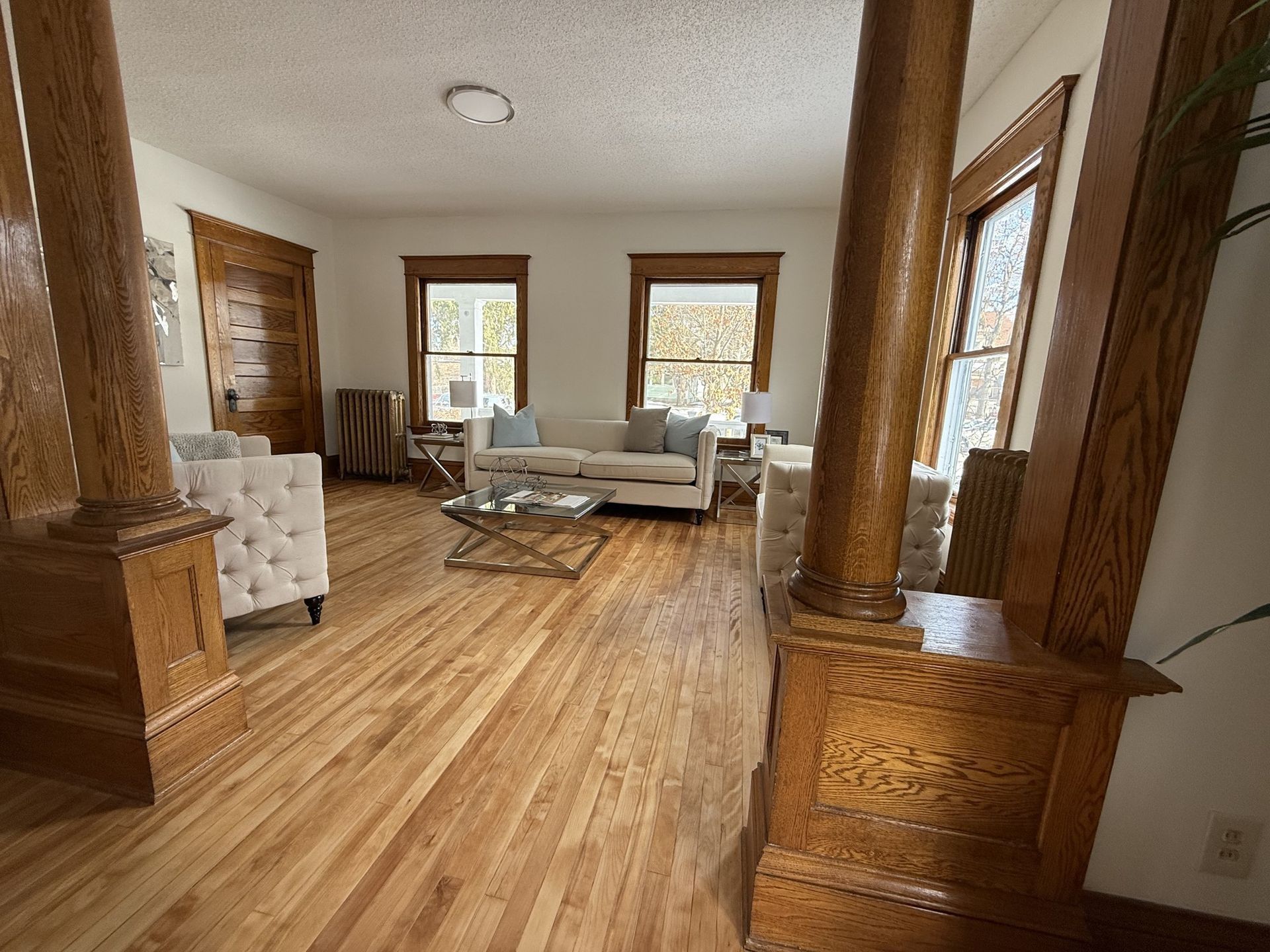 Spacious living room with hardwood floors, white sofas, large windows, and carved wooden columns