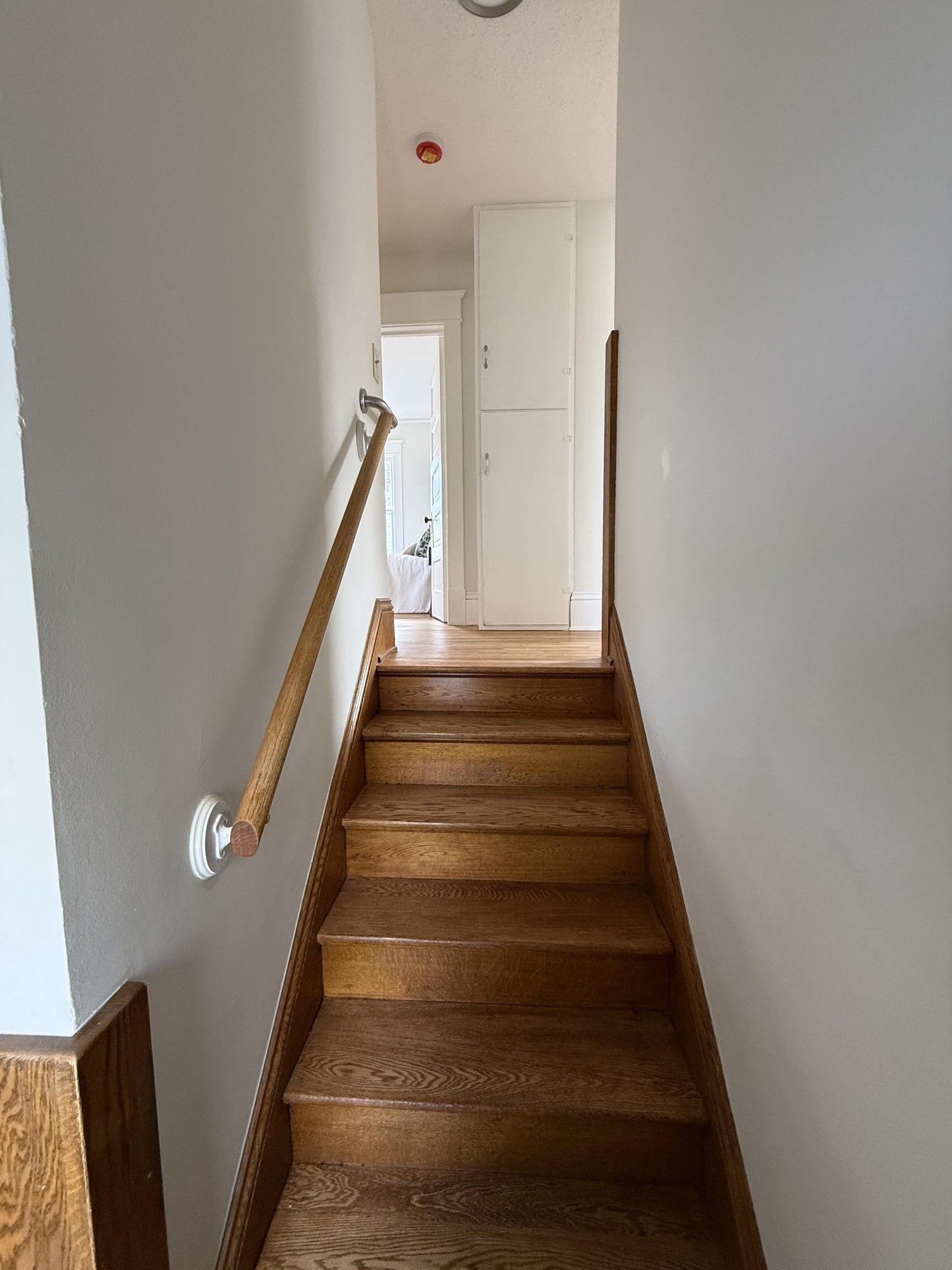 Narrow indoor stairway with wooden steps, white walls, and a handrail leading to a hallway.