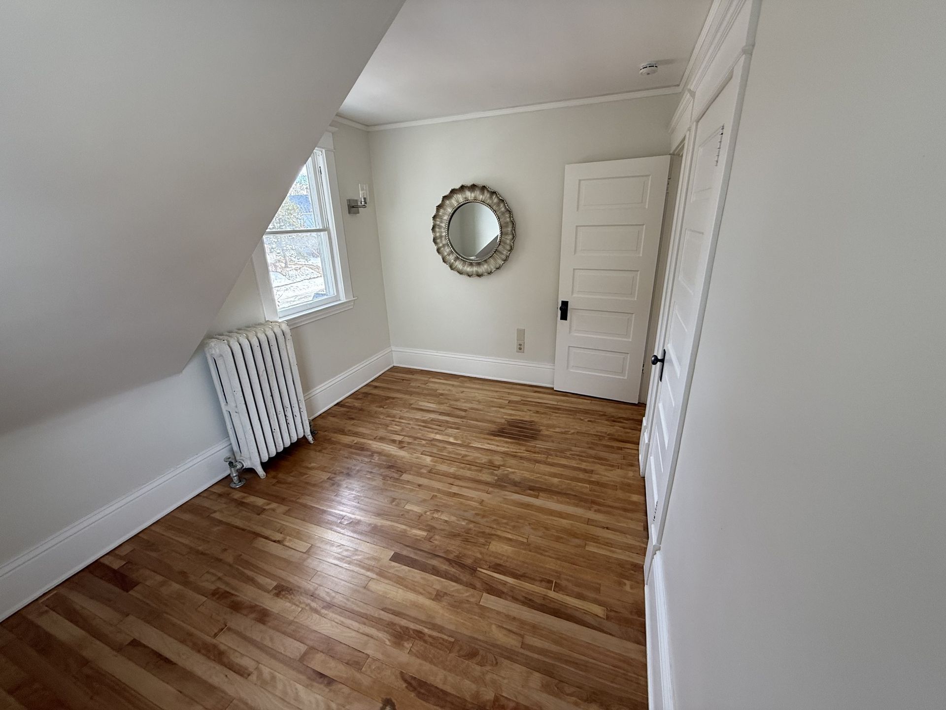 Small empty attic room with sloped ceiling, hardwood floor, window, radiator, and round mirror on the wall