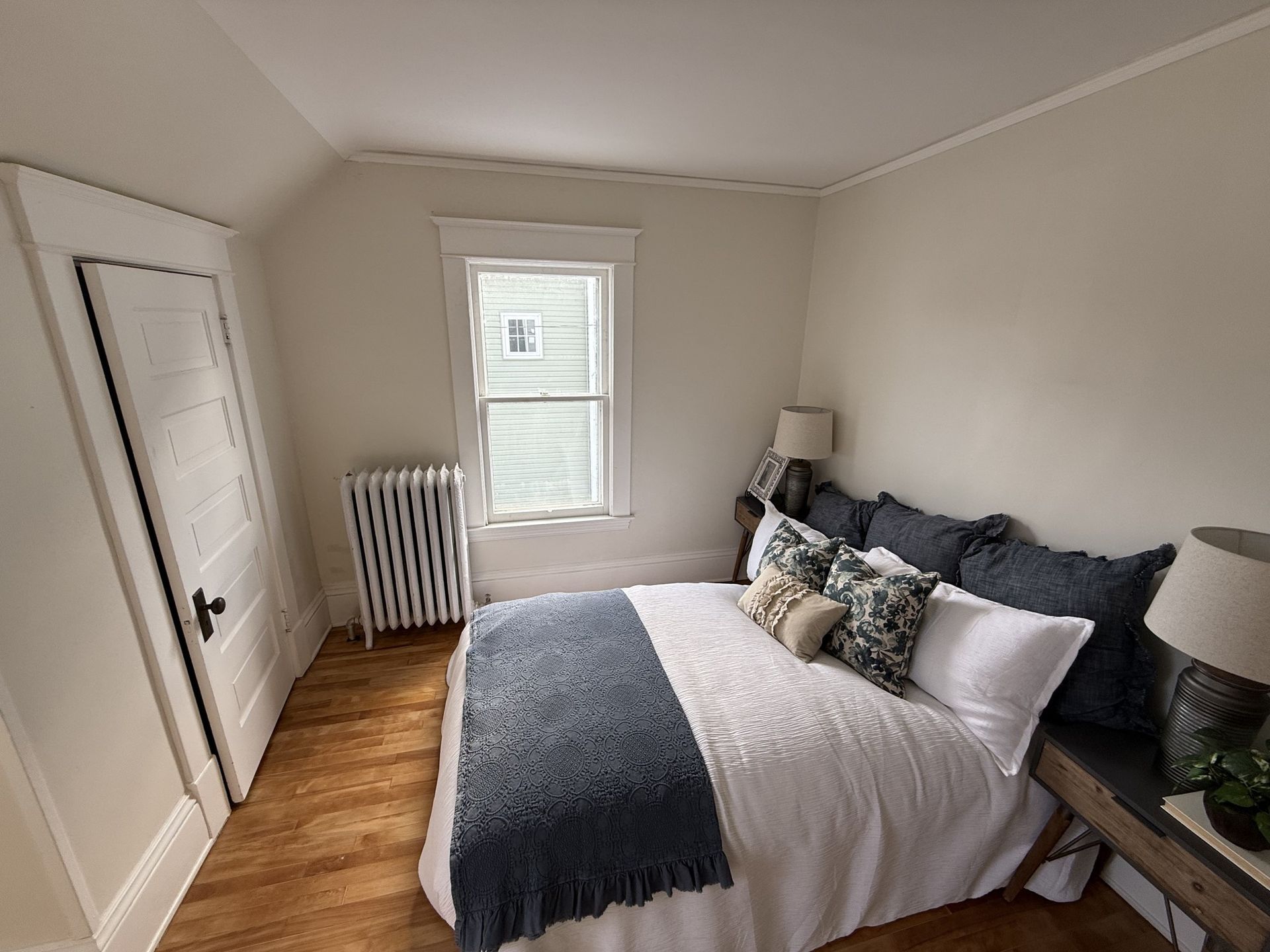 Bright bedroom with a bed, window, radiator, and white closet doors on wood floors.