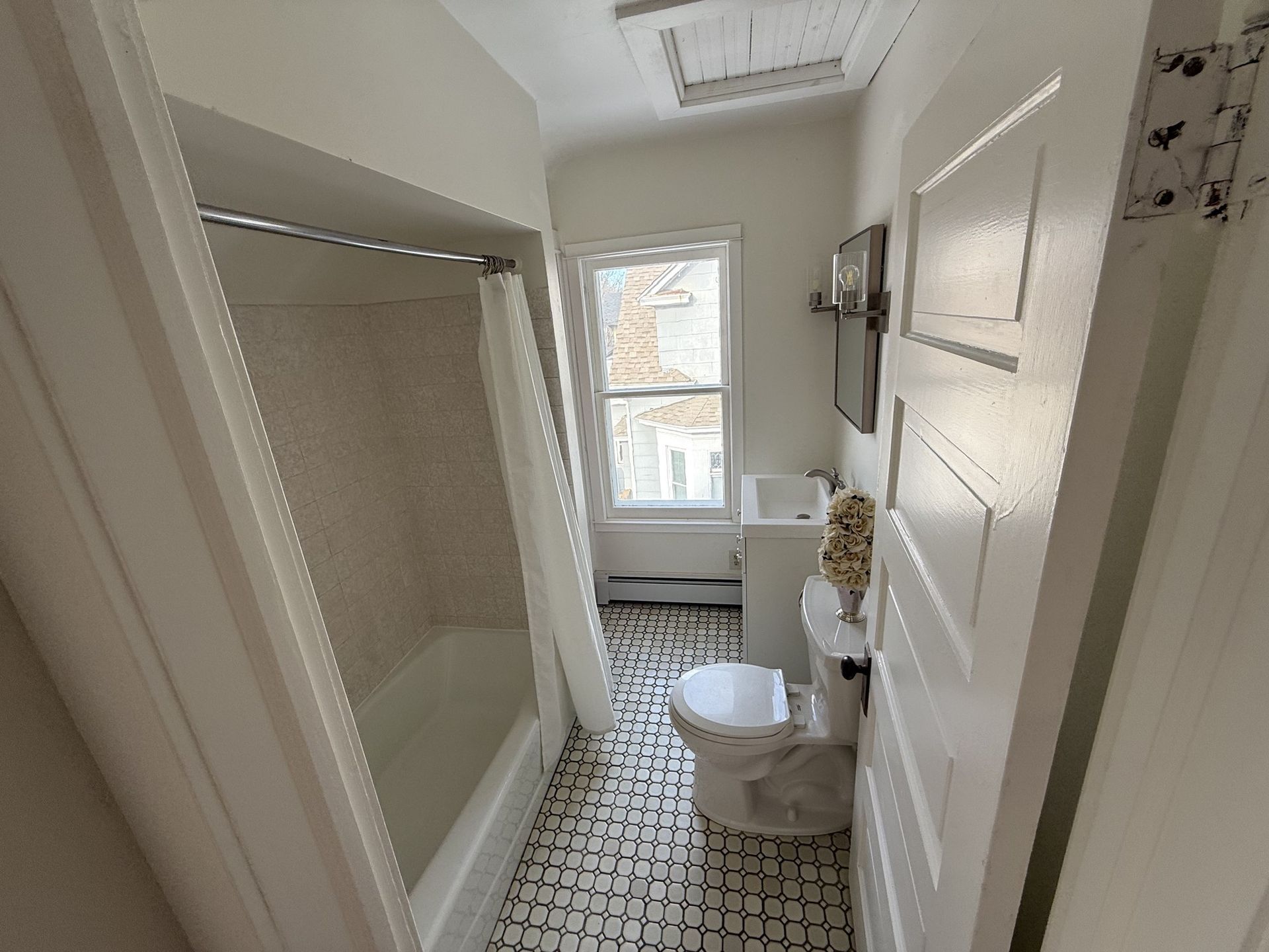 Narrow bathroom with white tub, toilet, sink, and window on patterned tile floor.