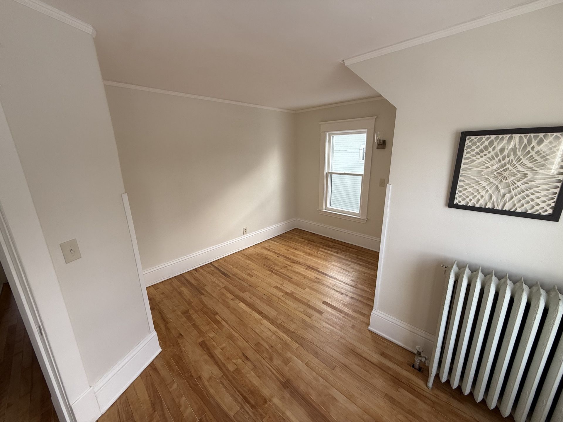 Empty room with hardwood floor, cream walls, white trim, radiator, and a small window.
