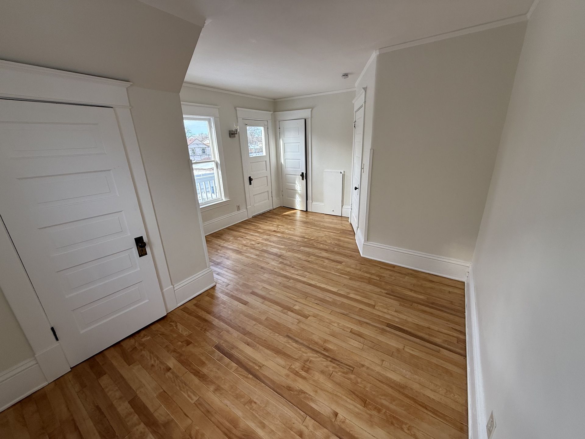 Empty beige room with hardwood floors, white trim, and a doorway leading to a brighter hall
