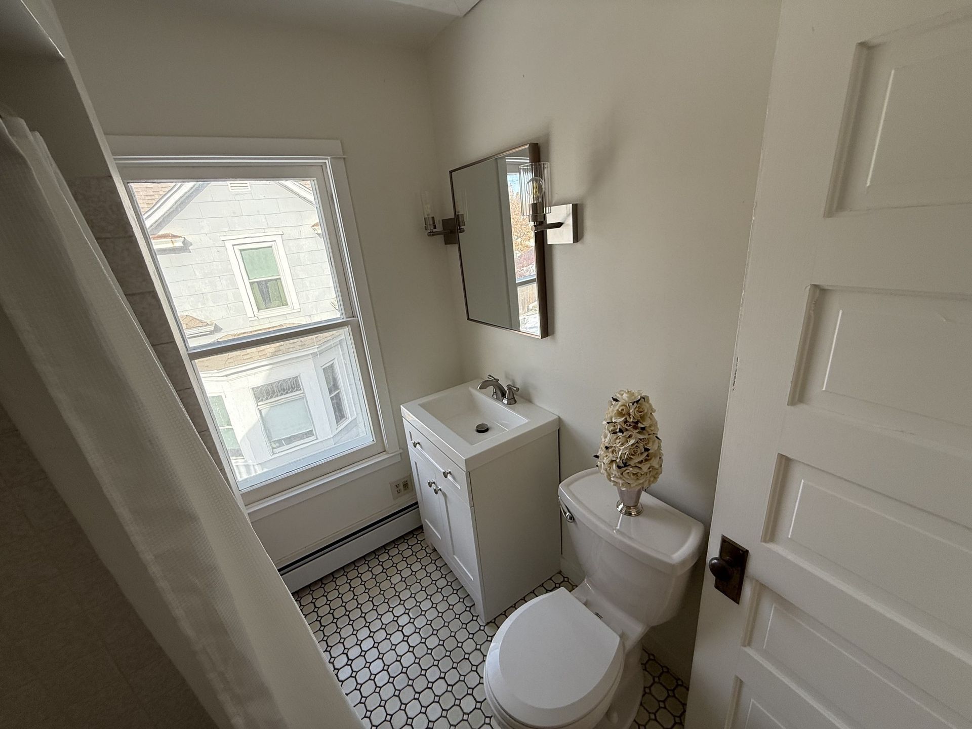 Small bathroom with toilet, vanity, mirror, and window over mosaic tile floor.