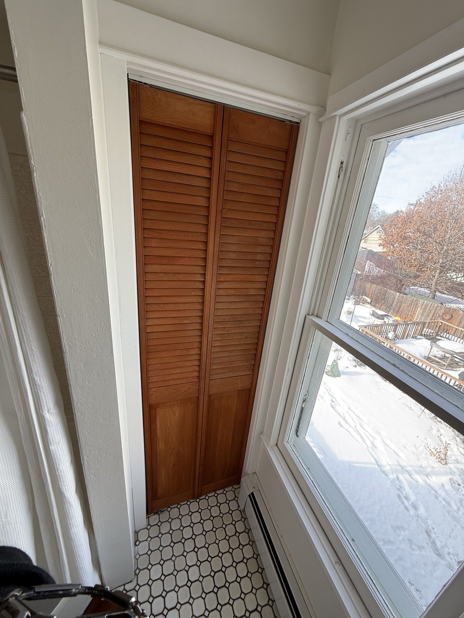 Narrow hallway closet with brown louvered doors beside a bright window and patterned tile floor