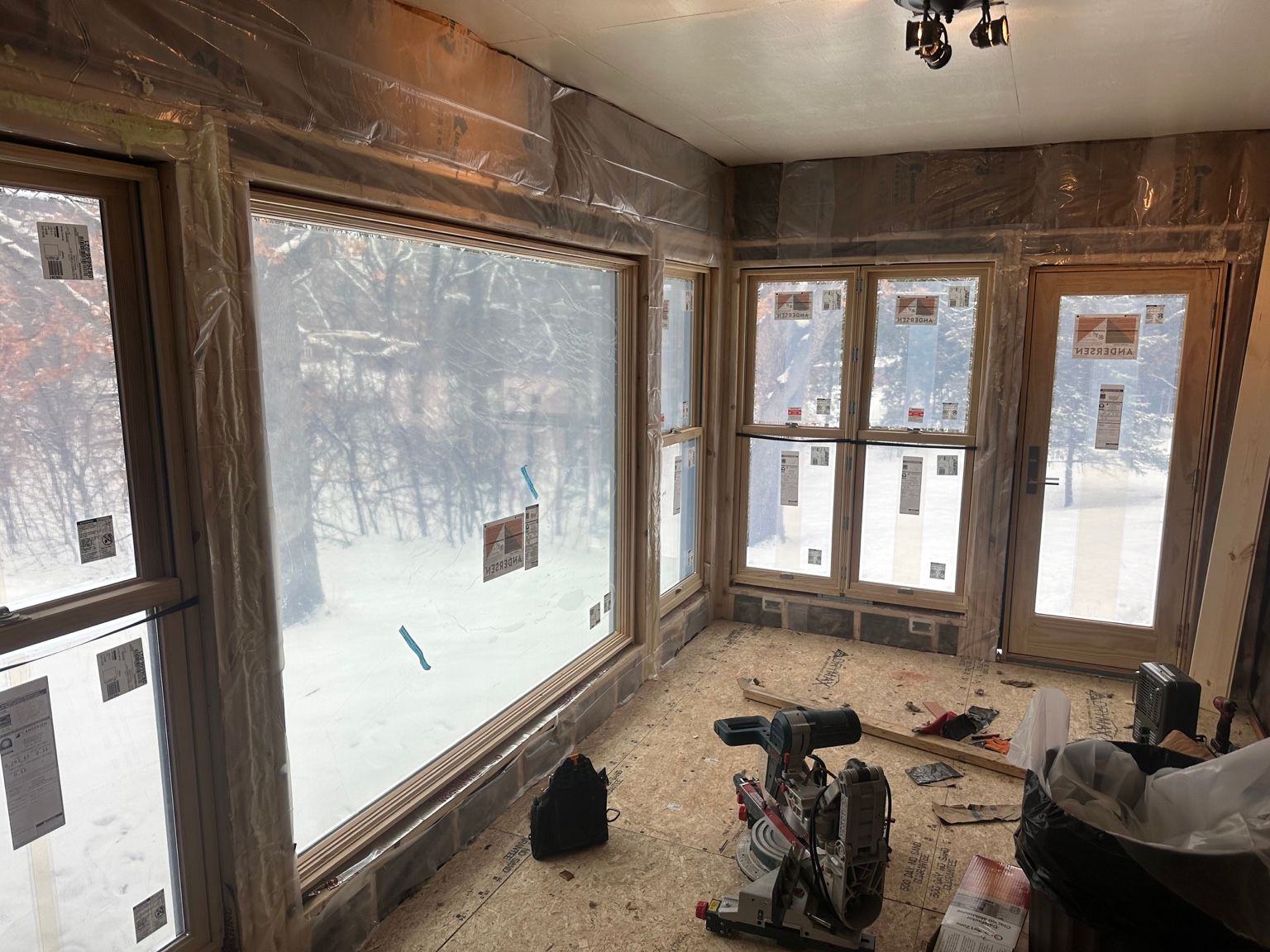 Sunlit unfinished room with exposed wood walls, large windows, and construction tools on the floor.