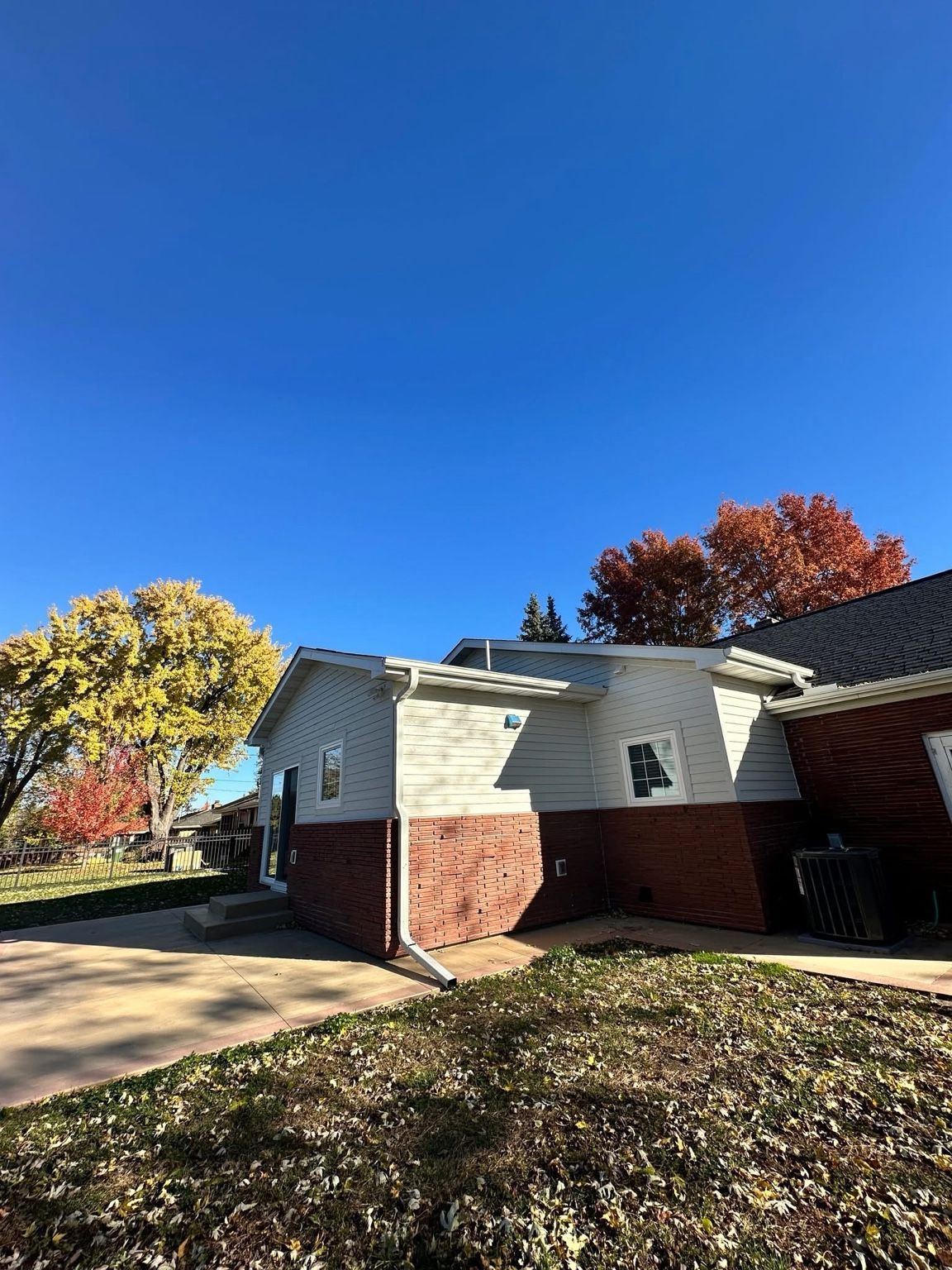 Suburban house with white siding and red brick under a bright blue sky, framed by autumn trees