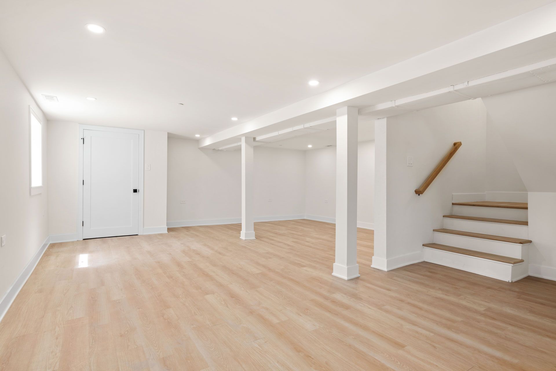 Bright empty basement room with white walls, wood floor, and stairs on the right