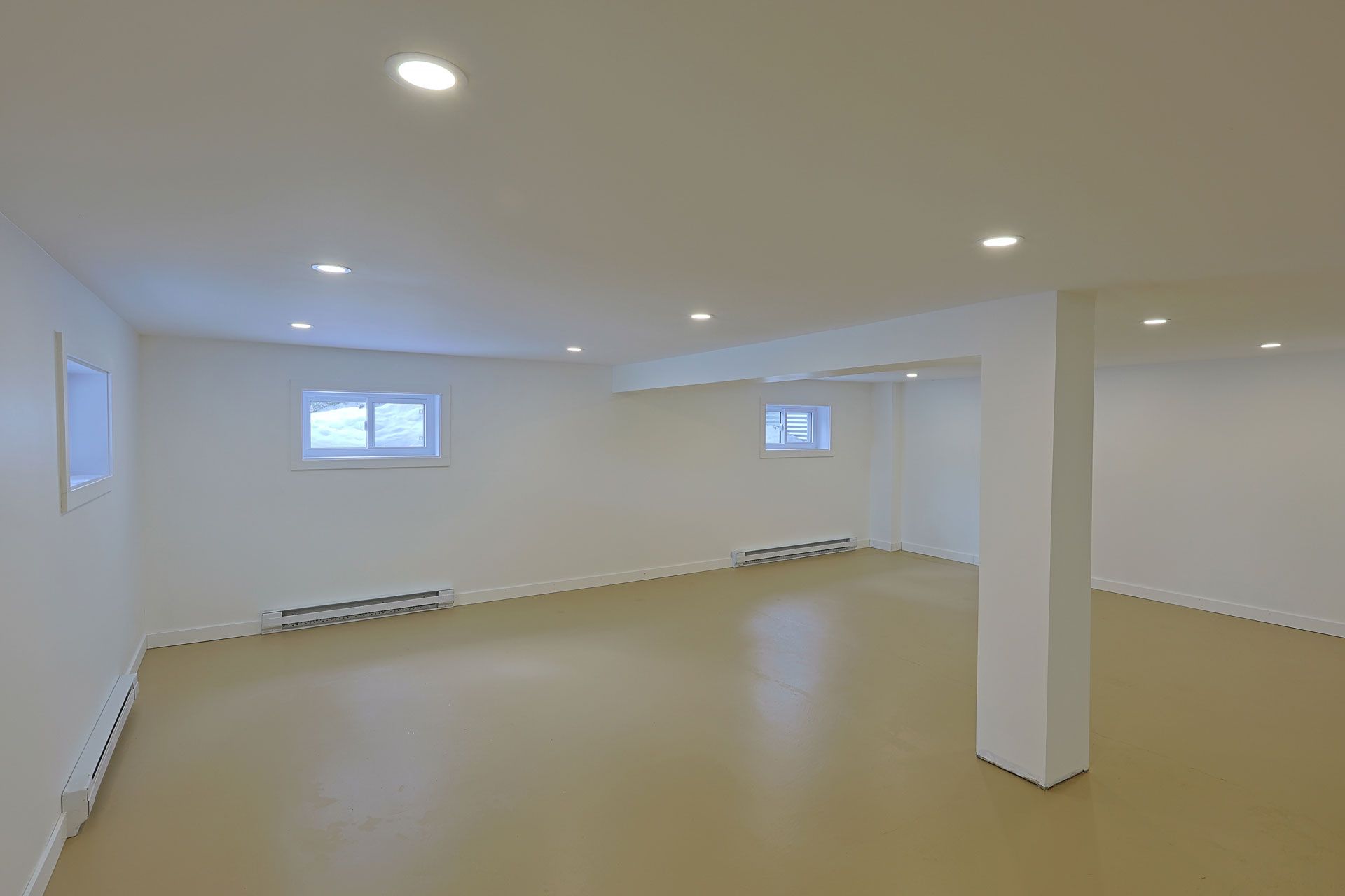 Empty white basement room with beige floor, recessed lights, windows, and a support column