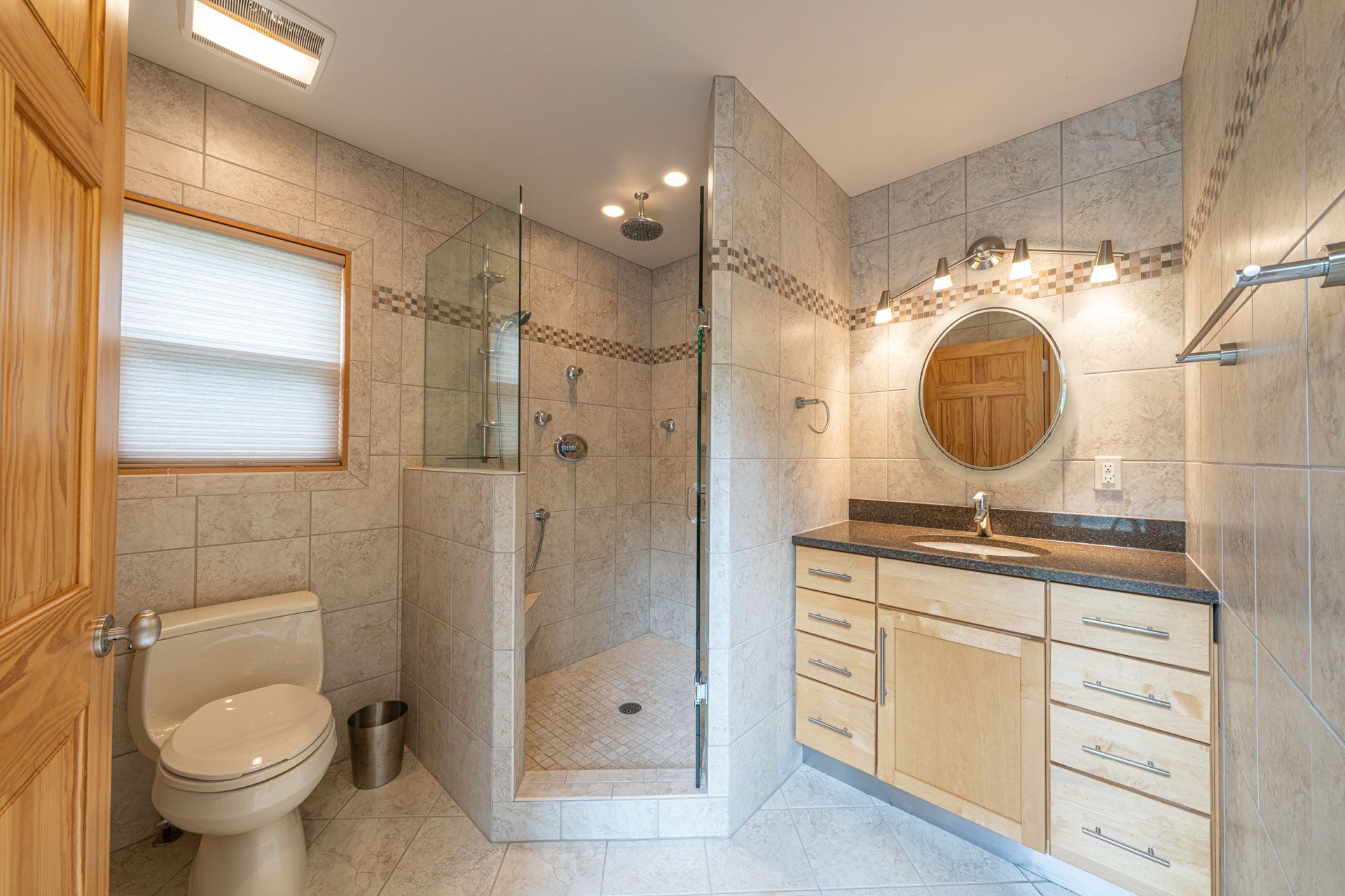 Bright bathroom with shower, toilet, vanity, and round mirror, in beige tile and wood accents