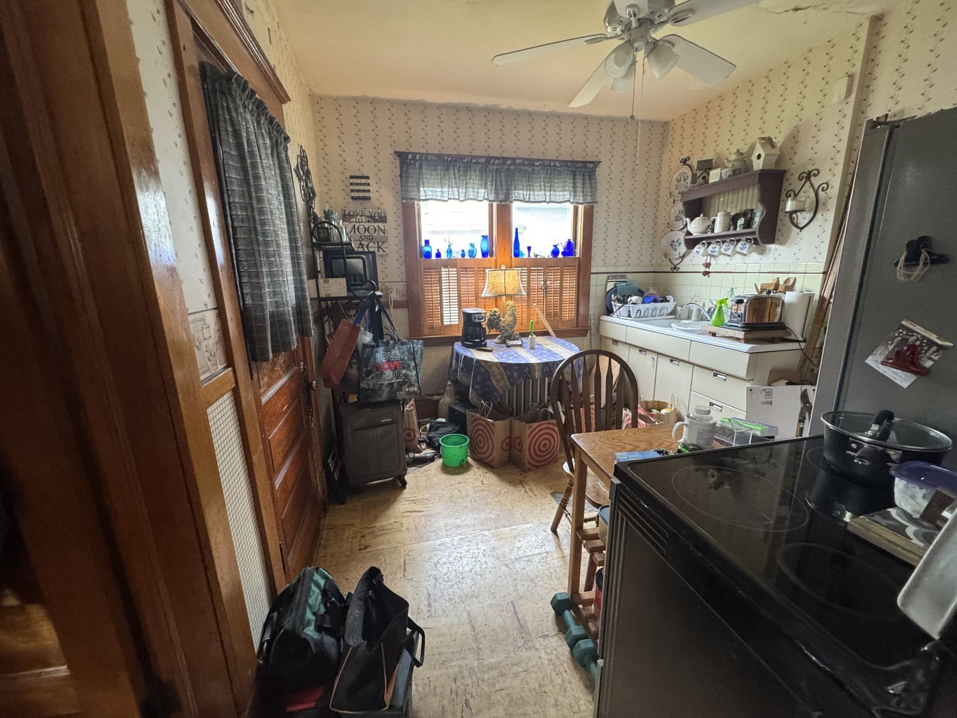 Cluttered kitchen with table, chairs, sink, stove, and a window with blinds and plants.