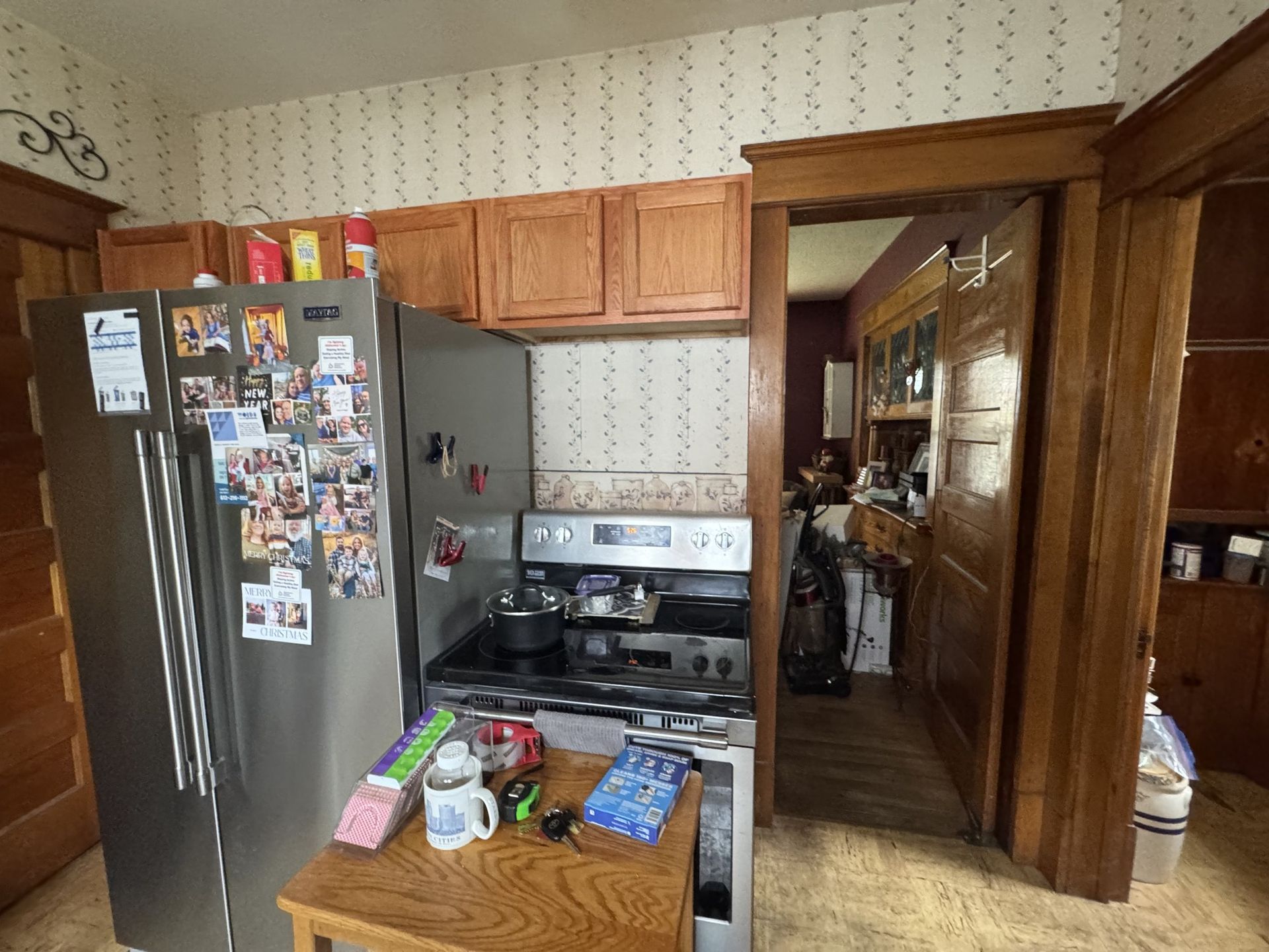 Small kitchen with wooden cabinets, stainless fridge covered in photos, and a cluttered counter beside the stove.