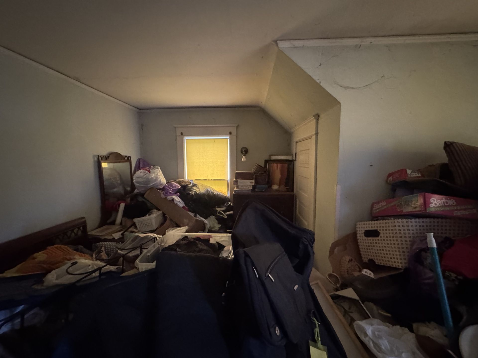 Cluttered bedroom with piles of clothes and belongings around a bed, window glowing at the back.