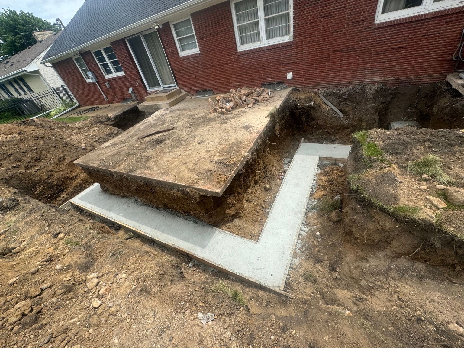 Foundation excavation and house leveling with exposed concrete footing beside a red brick home