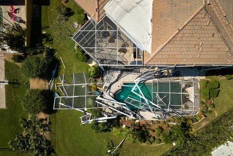 Collapsed metal pool cage beside a house, with a damaged teal pool area and debris in the yard.