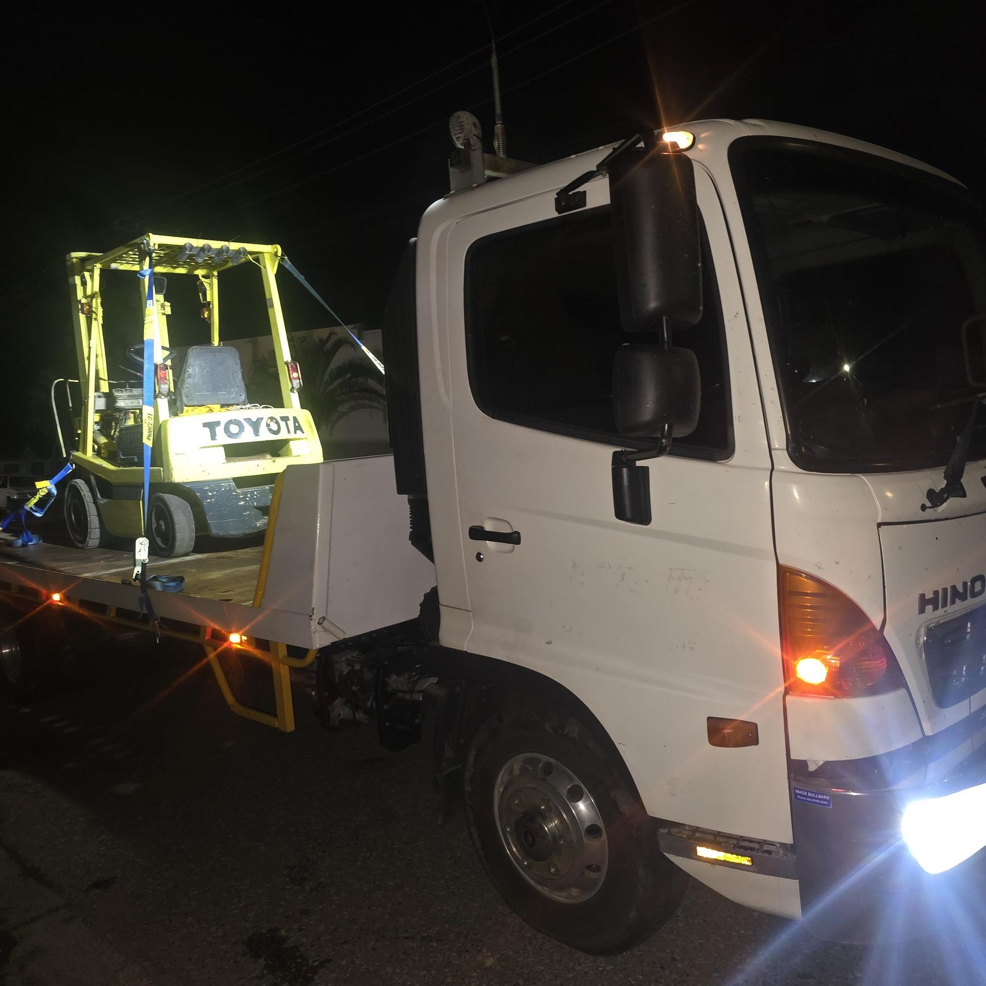 A White Tow Truck at Night Transporting a Yellow Toyota Forklift — Old Mate's Towing Townsville in Deeragun, QLD